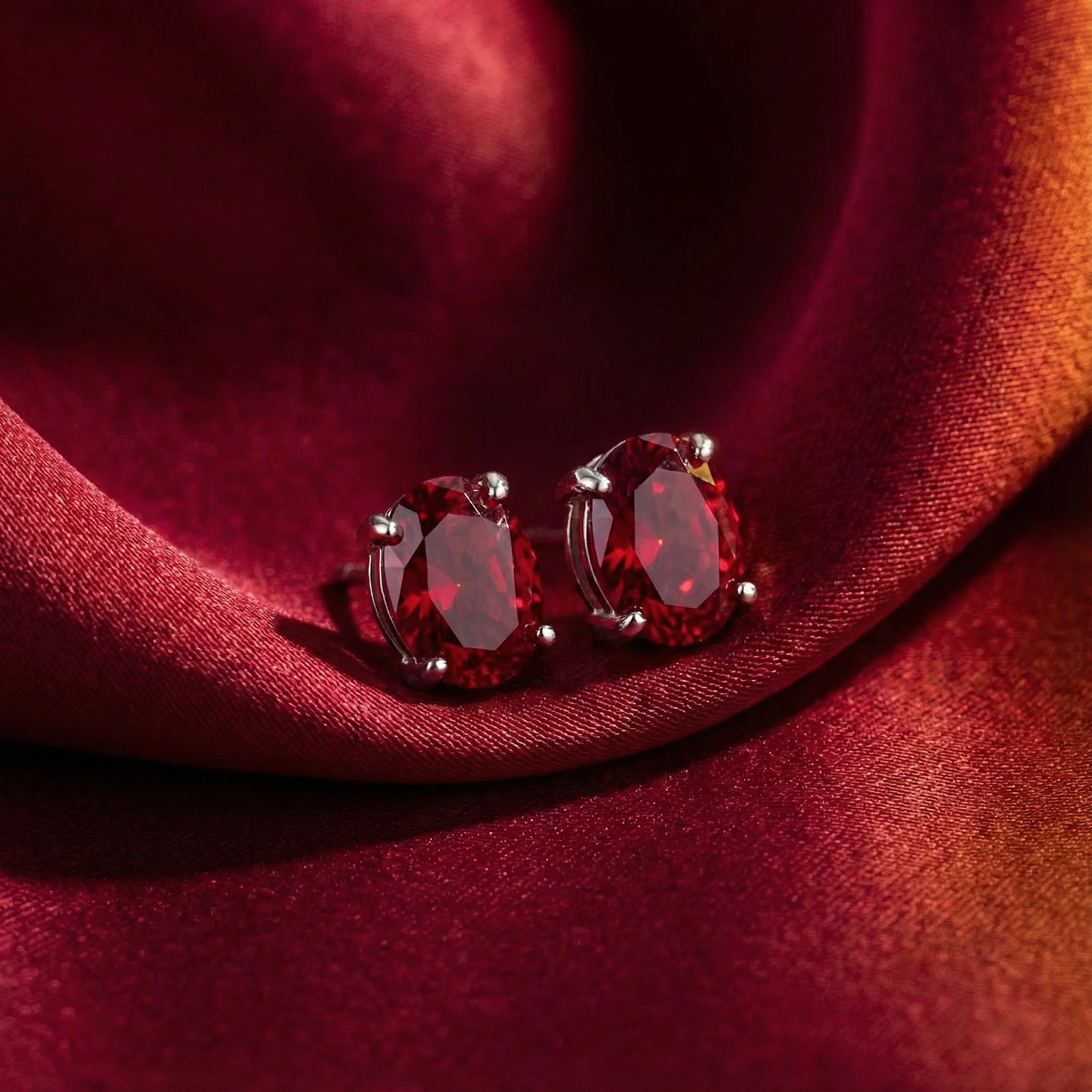 Oval-cut simulated ruby stud earrings in sterling silver resting on draped red satin fabric.