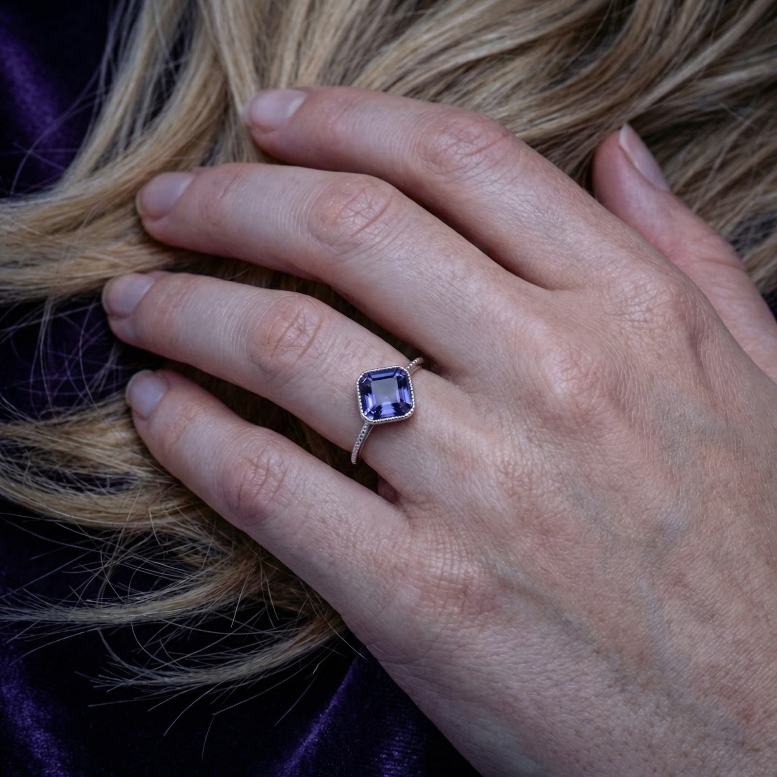 Square-cut simulated tanzanite silver ring worn on a woman's hand against purple velvet.