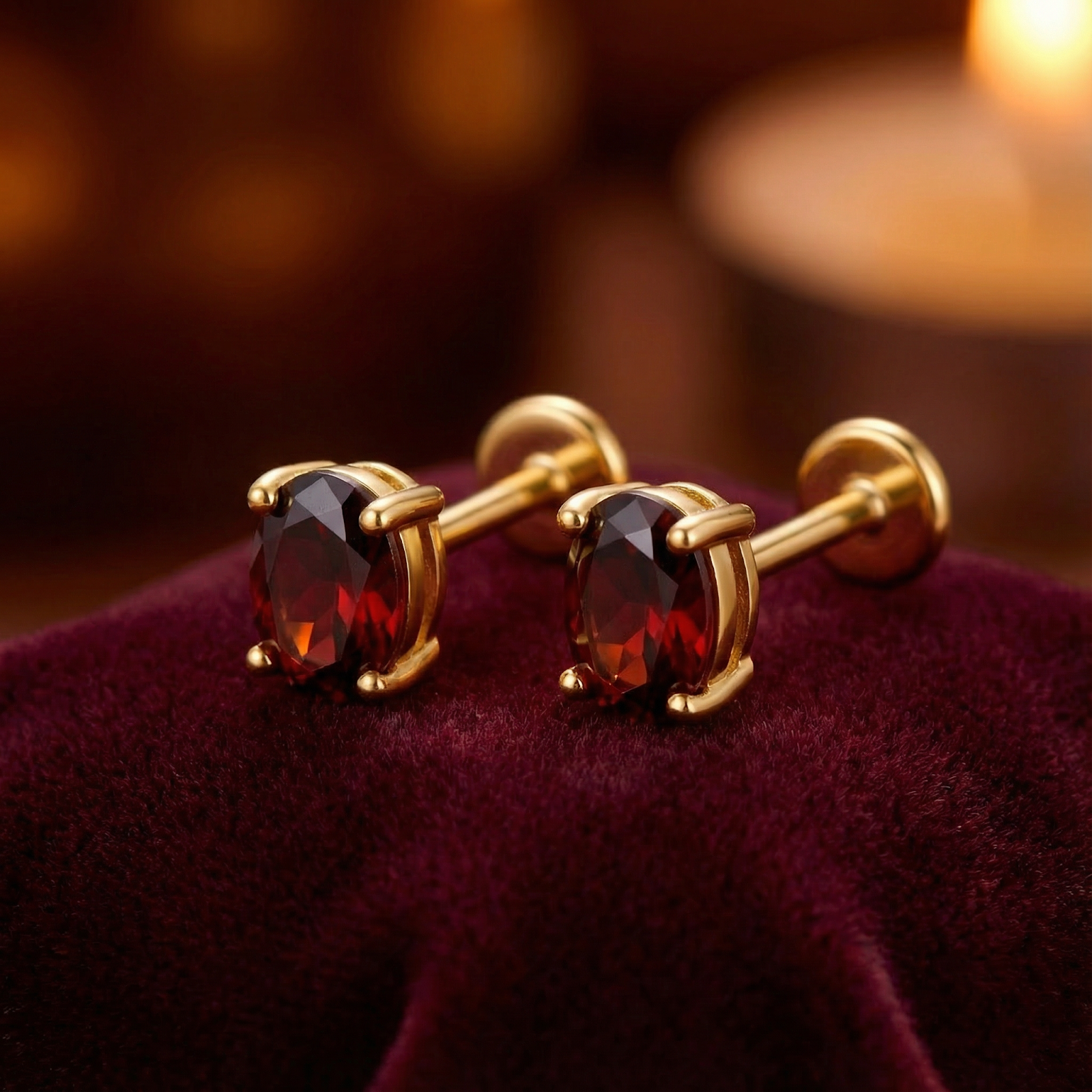 Oval cut simulated garnet stud earrings in 18K gold vermeil resting on red velvet with warm background lighting.