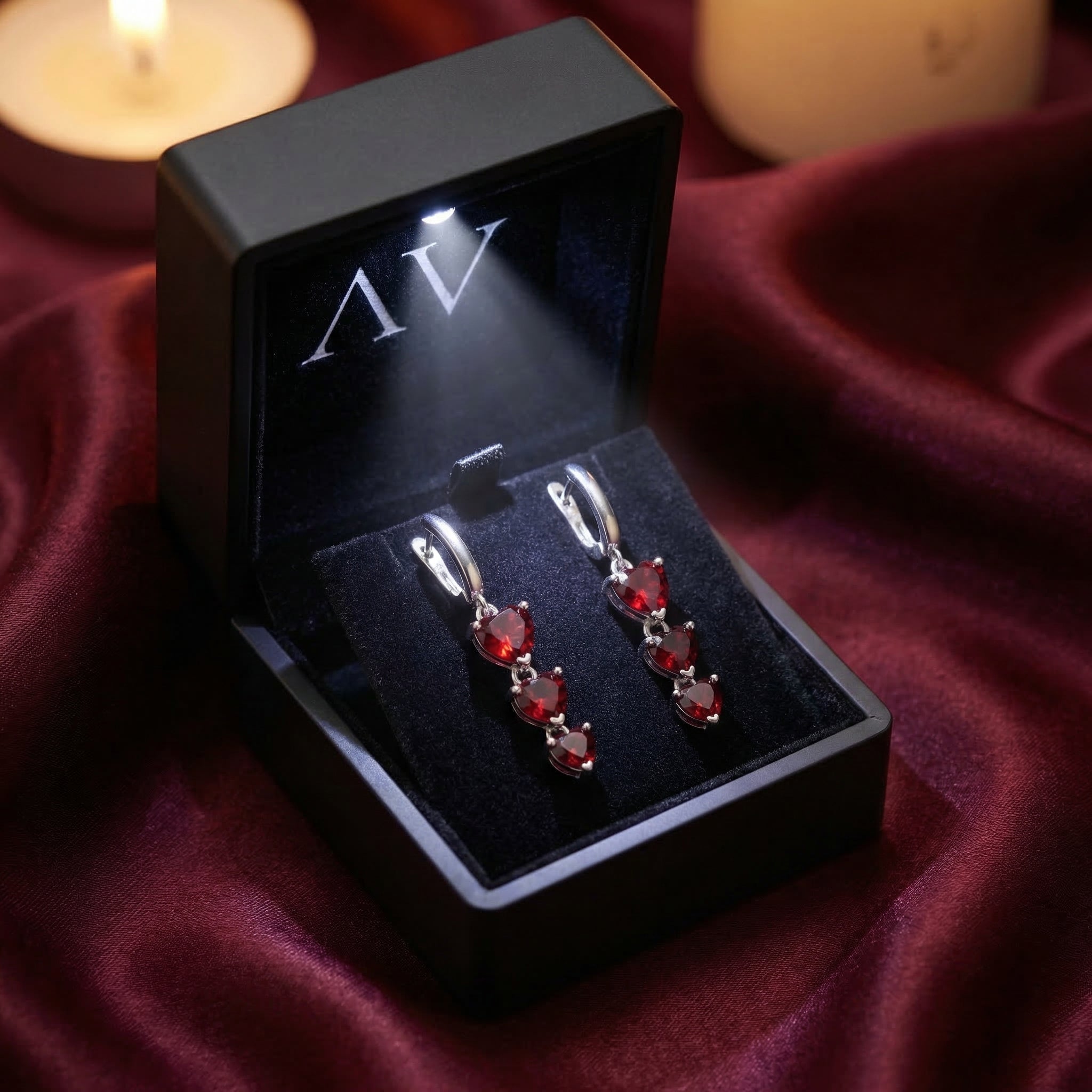 Silver heart drop earrings with red garnets inside a black LED-lit gift box, resting on burgundy silk fabric.