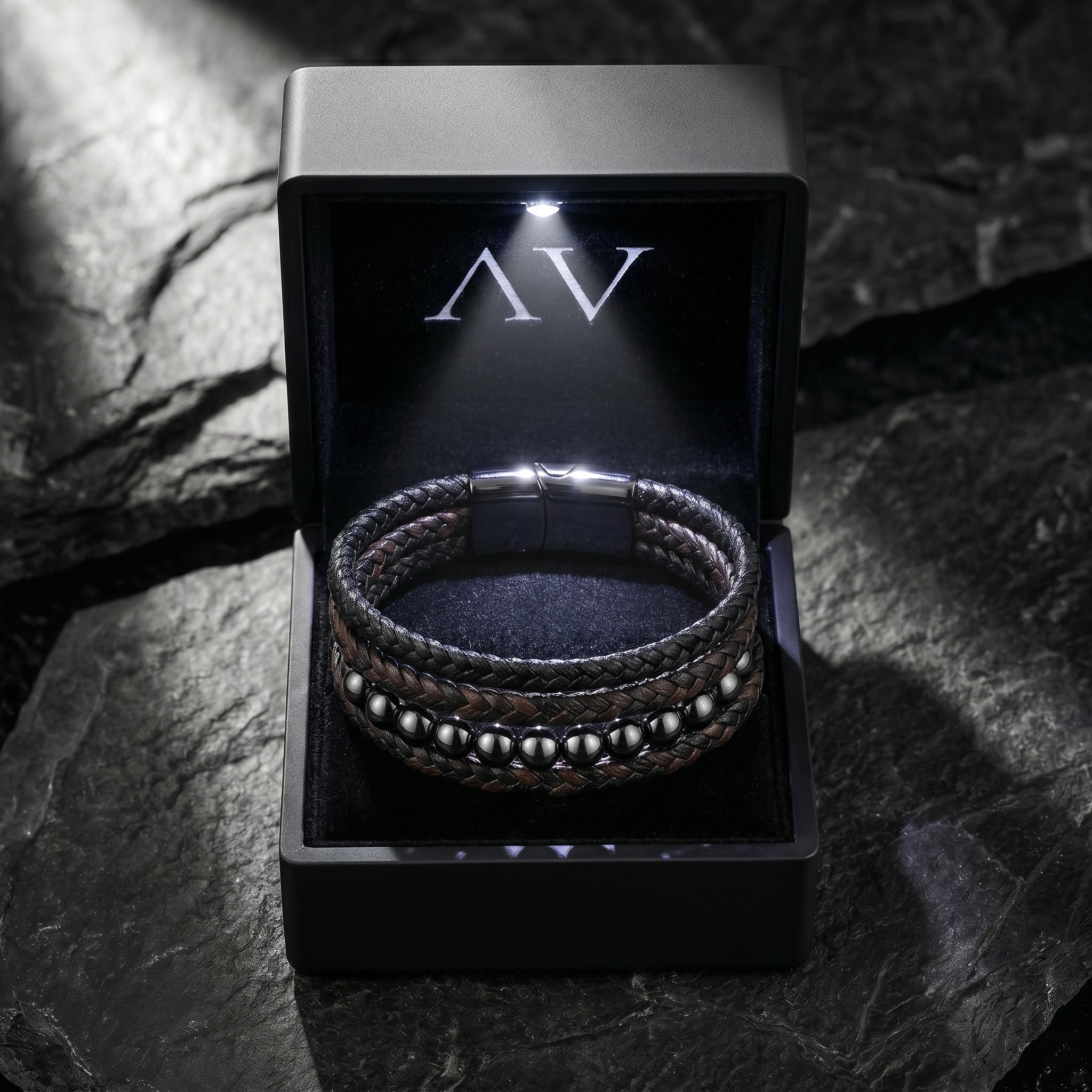 Men's leather and gunmetal bead bracelet in a black LED-lit branded gift box on a textured stone surface.