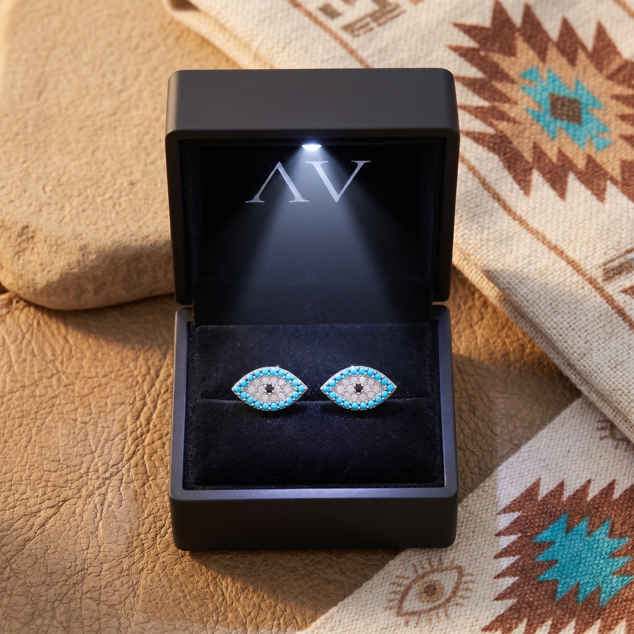 Silver turquoise evil eye stud earrings presented in a black LED-lit jewelry gift box on a patterned cloth.