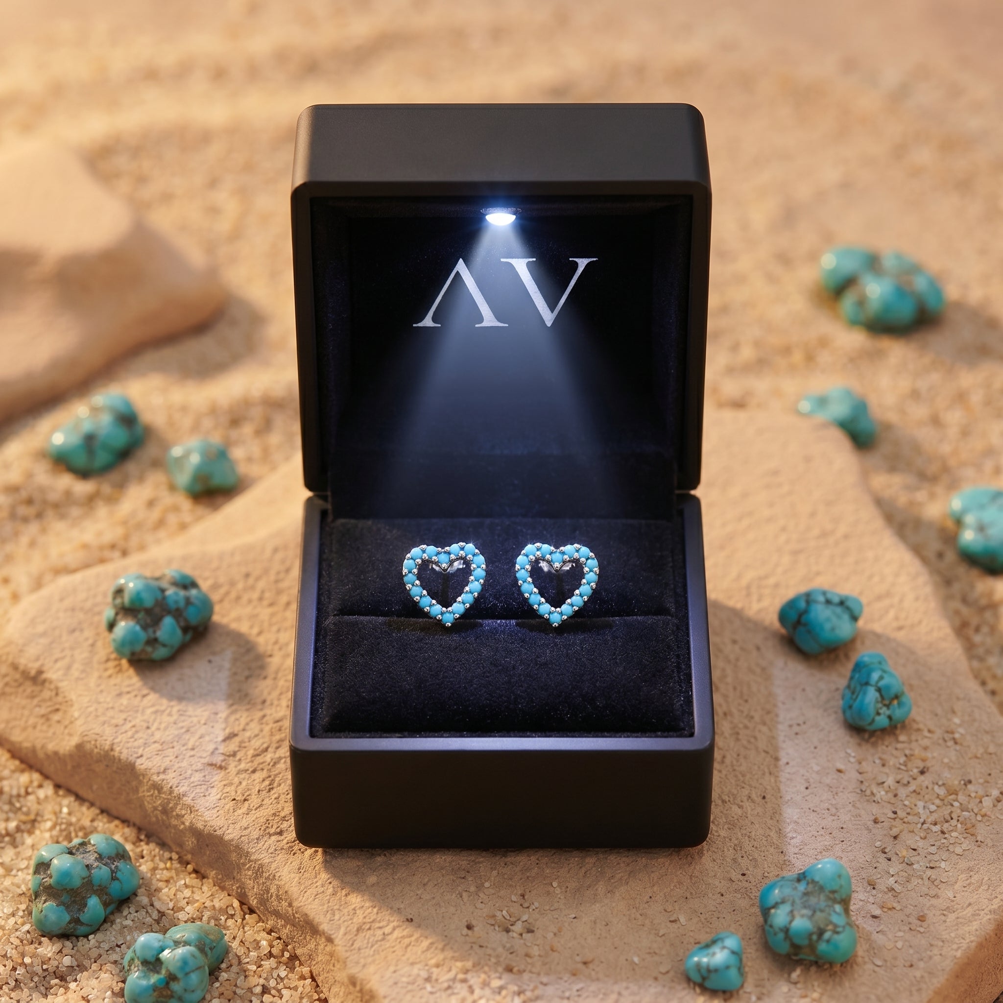 Silver heart-shaped turquoise earrings in a black LED-lit gift box on sand with raw turquoise accents.