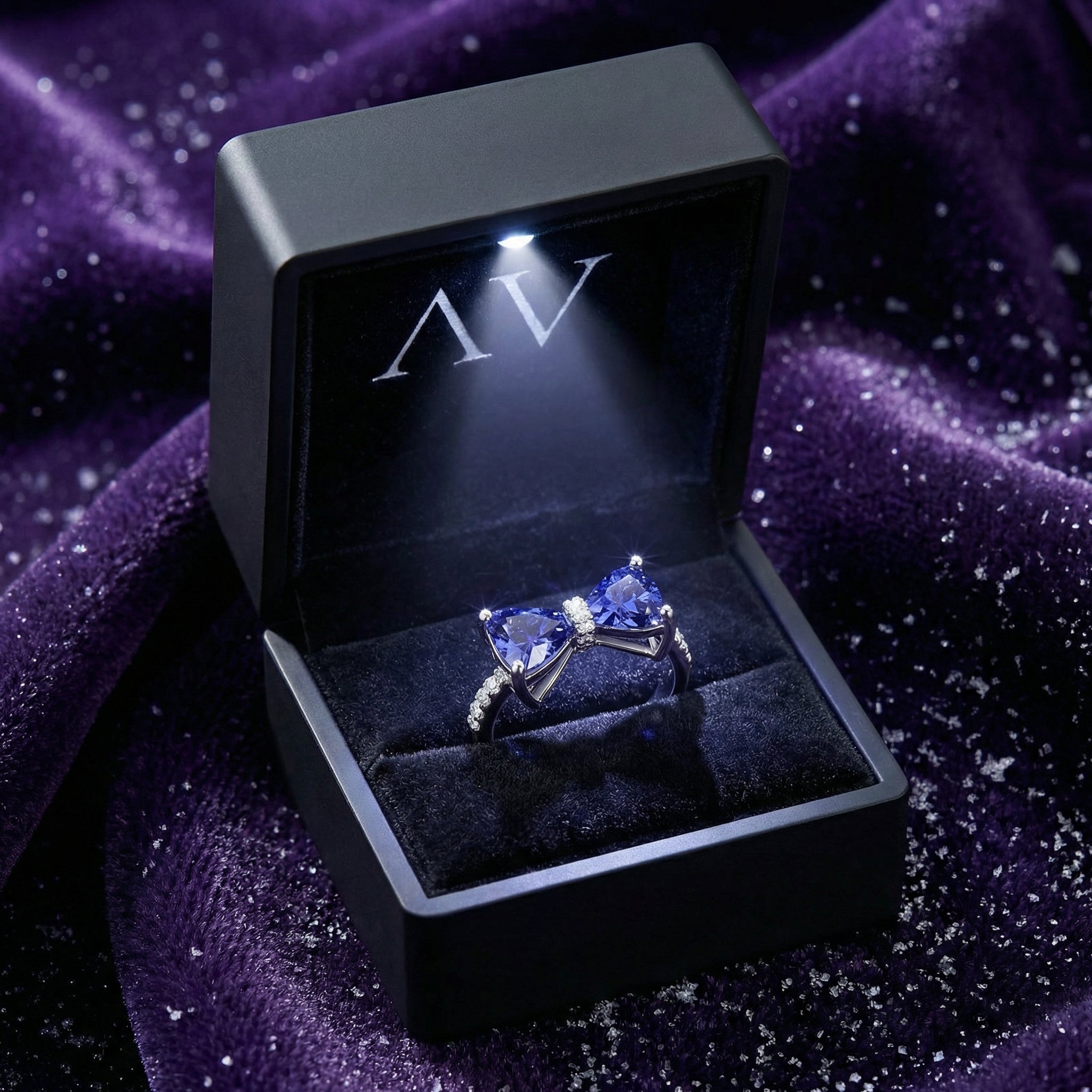 Evelia bow ring with simulated tanzanite stones inside a lighted black jewelry gift box on purple velvet.