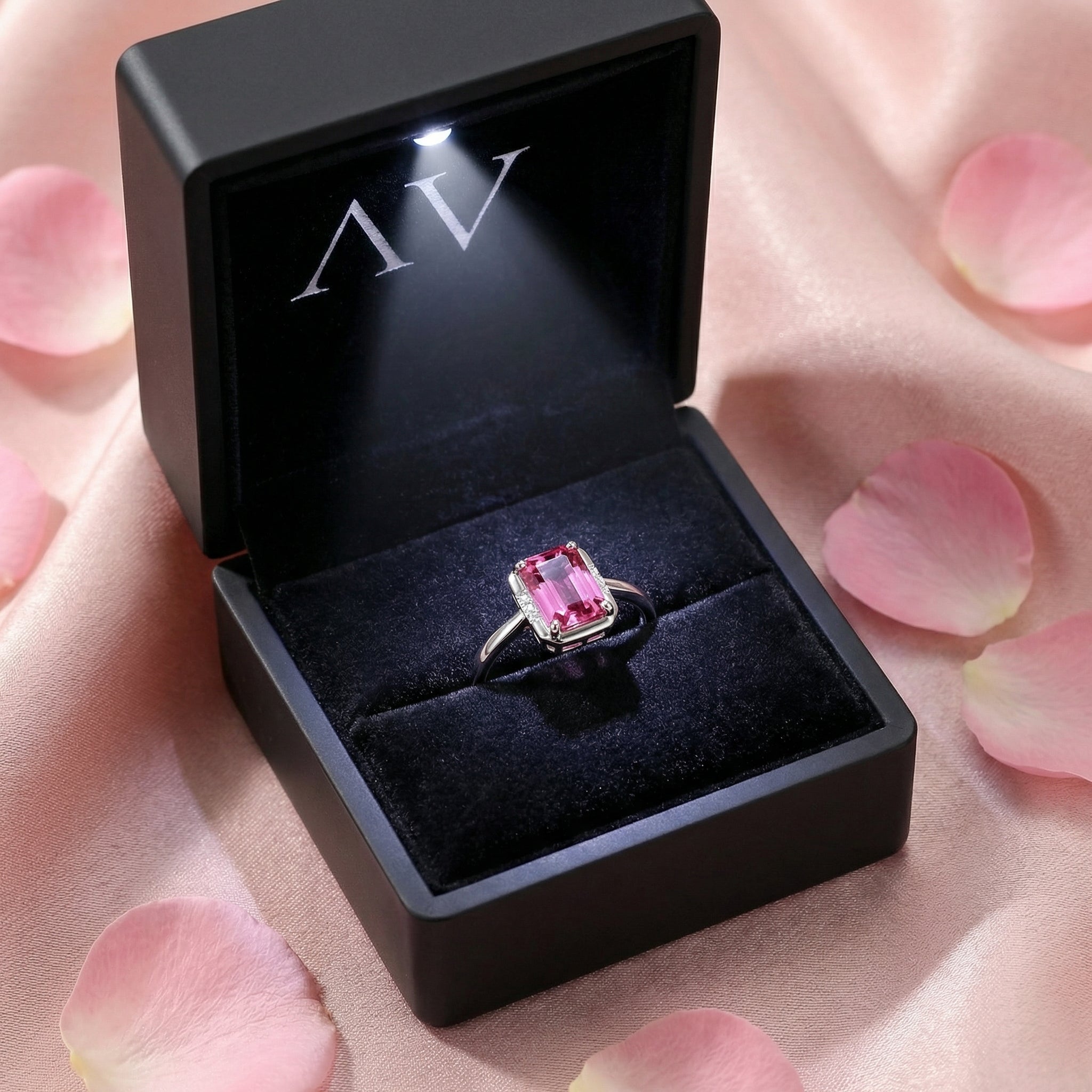 Emerald-cut pink tourmaline sterling silver ring inside a lighted black velvet jewelry box with rose petals.