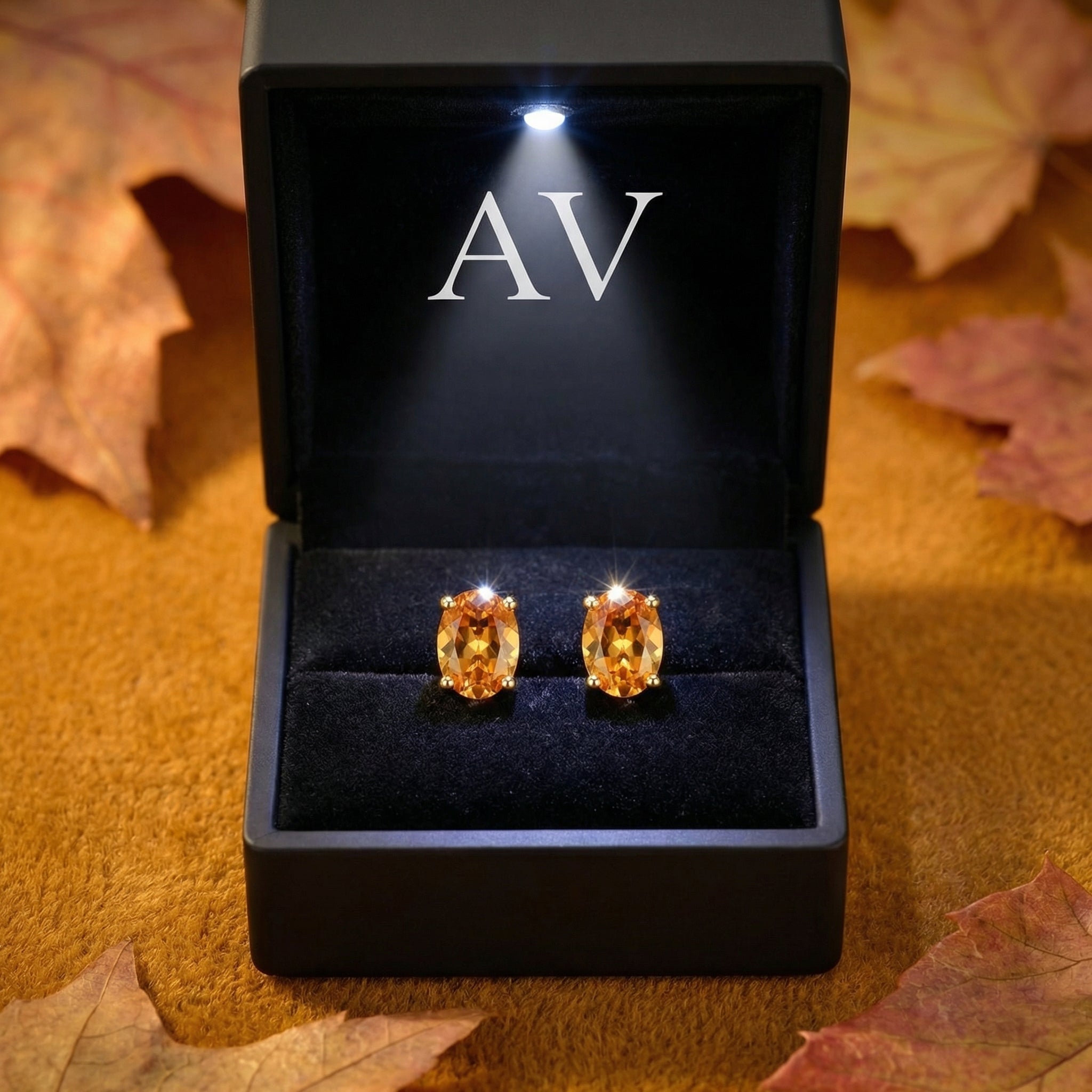 Oval simulated topaz stud earrings in 18K gold vermeil inside a lighted black jewelry box with leaves.