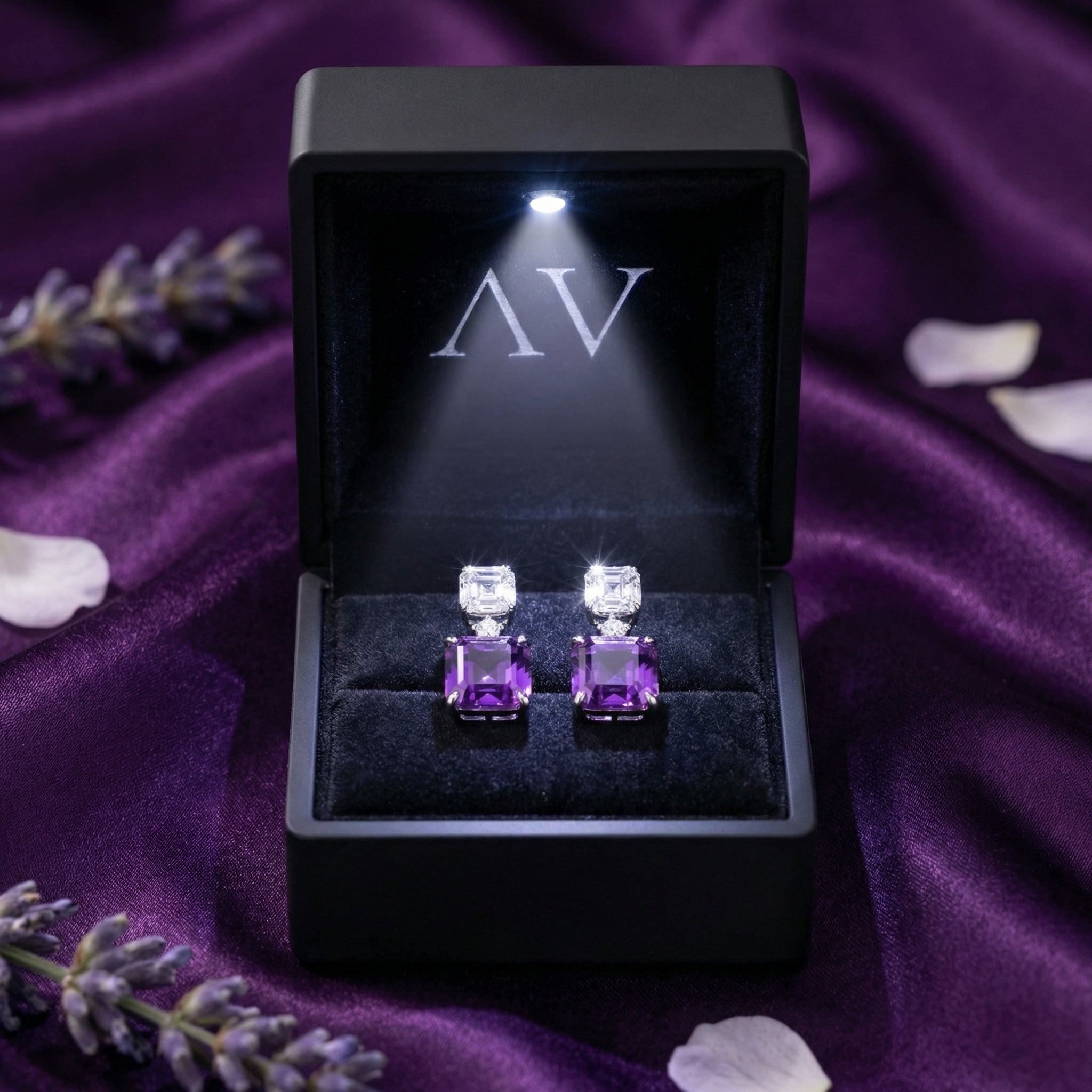 Pair of square cut amethyst drop earrings in a lighted black jewelry box on purple silk fabric.