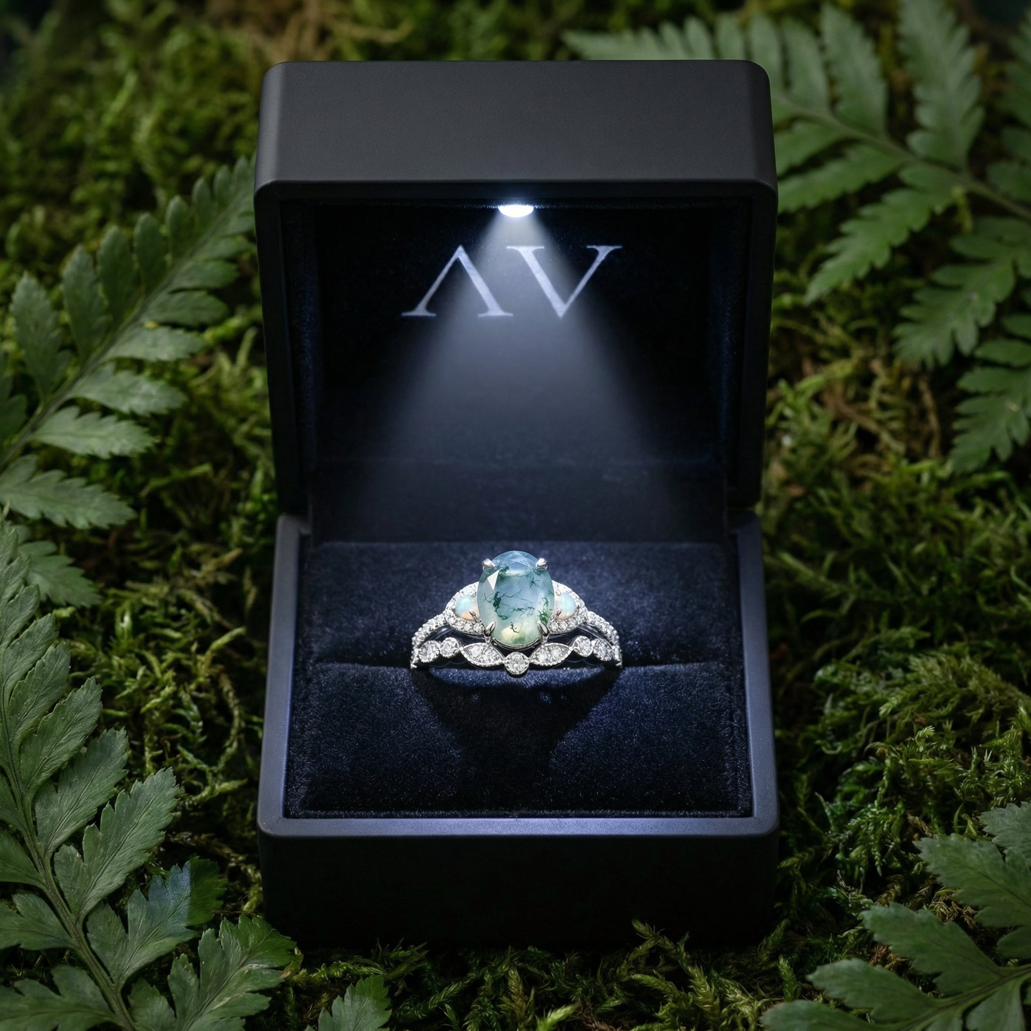 Oval cut moss agate ring set in a lighted black jewelry gift box surrounded by green ferns.