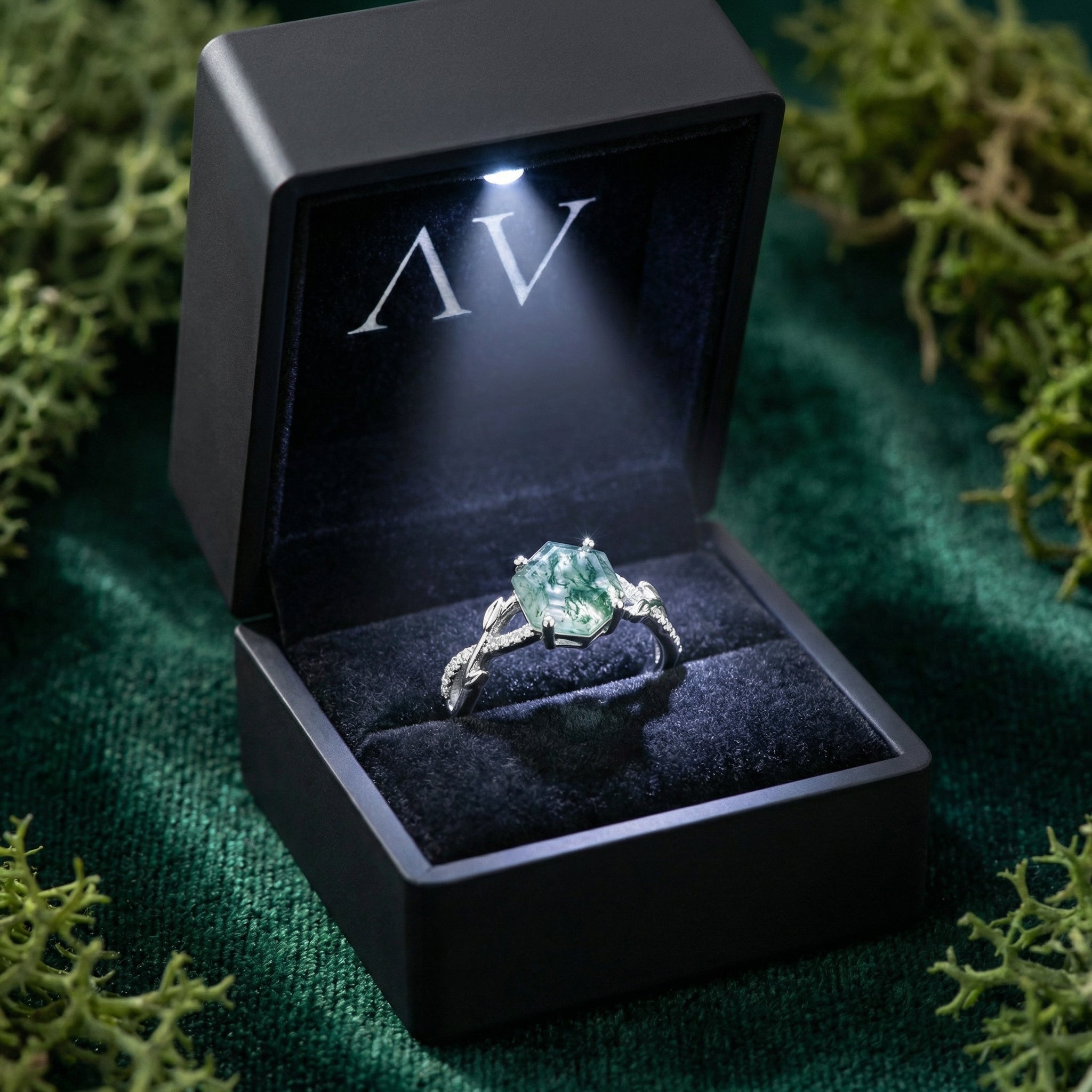 Hexagonal natural moss agate sterling silver ring in a black LED-lit gift box surrounded by green moss.