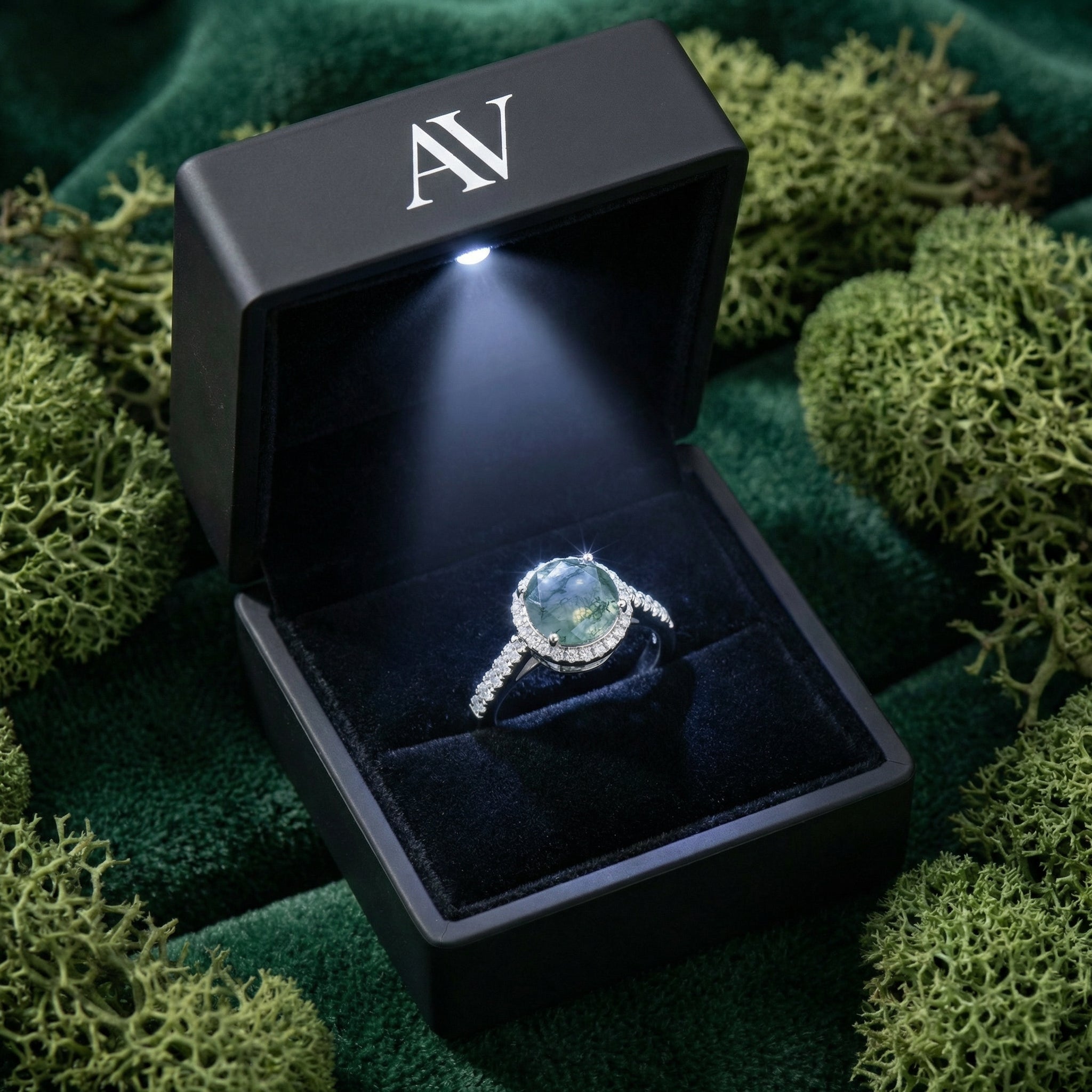 Natural moss agate ring in a black LED-lit gift box with AV logo, nestled in green moss.