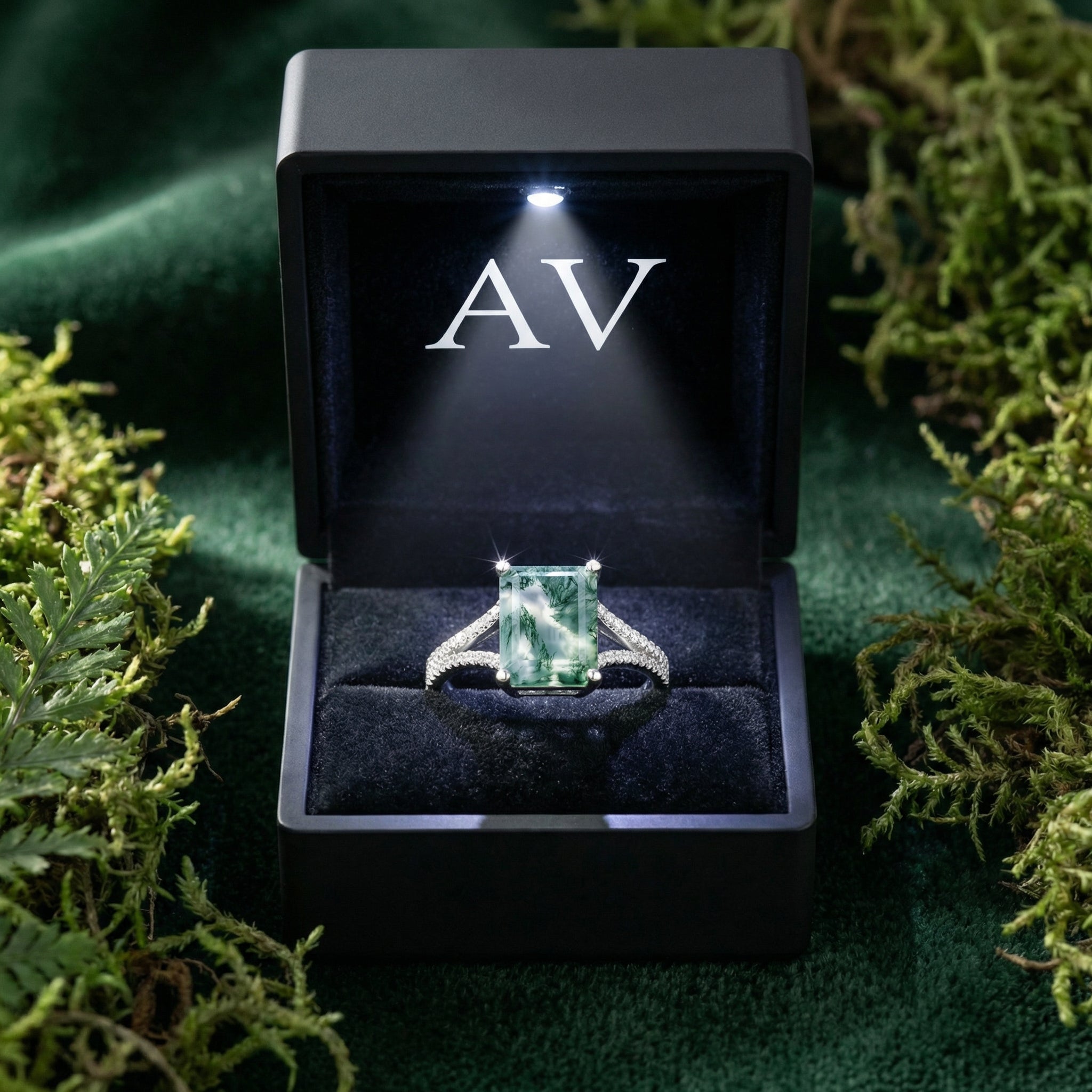 Cushion cut moss agate ring presented in a black LED-lit gift box with an AV logo, surrounded by green moss.