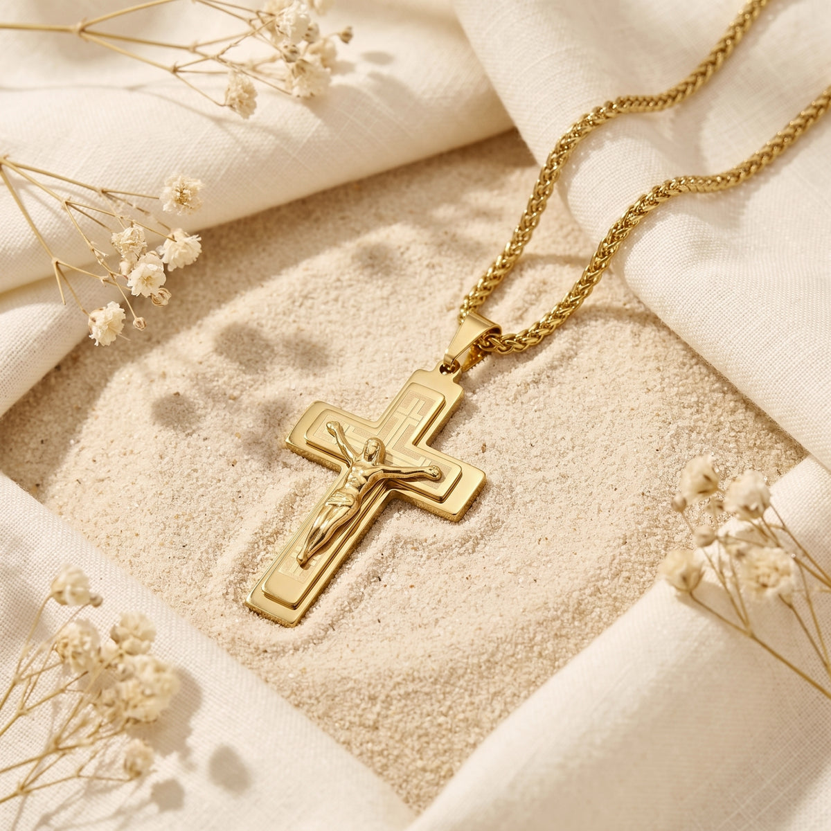 Gold-tone layered crucifix necklace resting on fine sand with white cloth and dried flowers