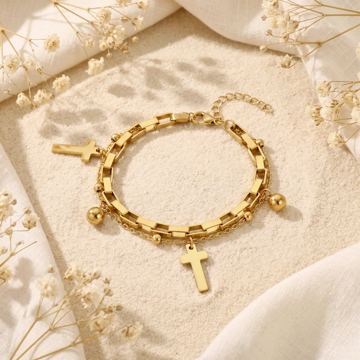 Gold double-layer bracelet with cross and sphere charms laid flat on sand with dried white flowers.