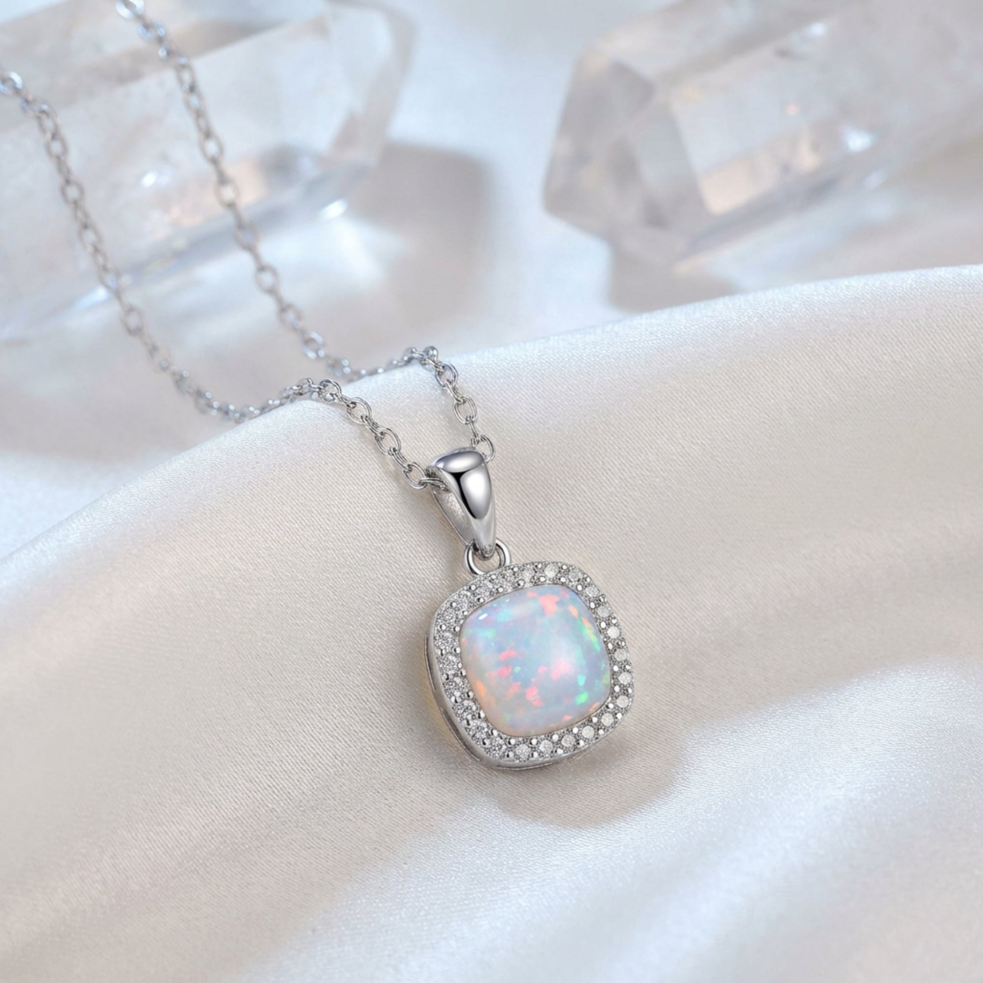 Silver cushion cut white opal halo pendant necklace on white silk fabric with decorative crystals.