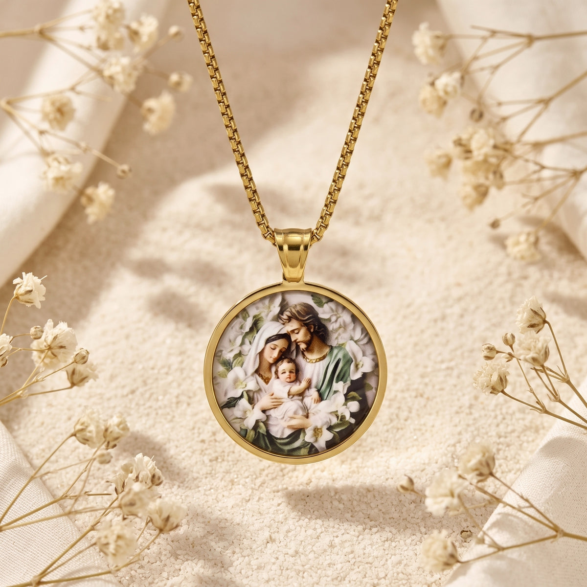 Gold-tone circular Holy Family medallion pendant on a box chain, displayed among dried white flowers.