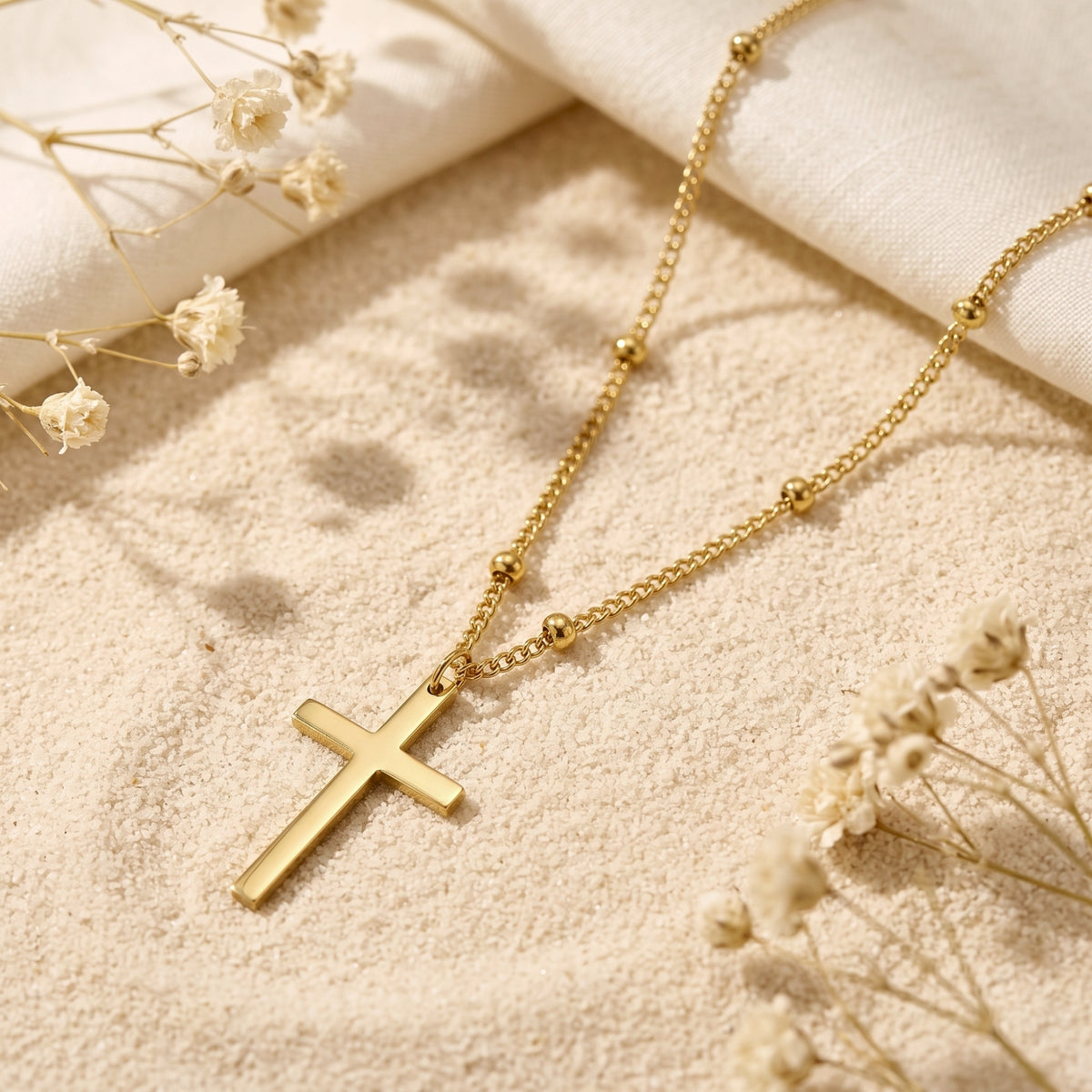 Gold-tone cross pendant and beaded station chain arranged on sand with dried white flowers.