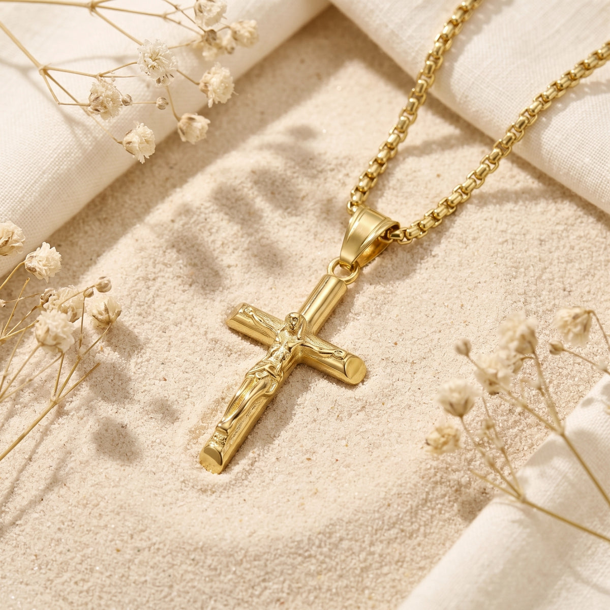 Gold-tone crucifix pendant resting at an angle on sand, surrounded by white fabric and dried flowers.