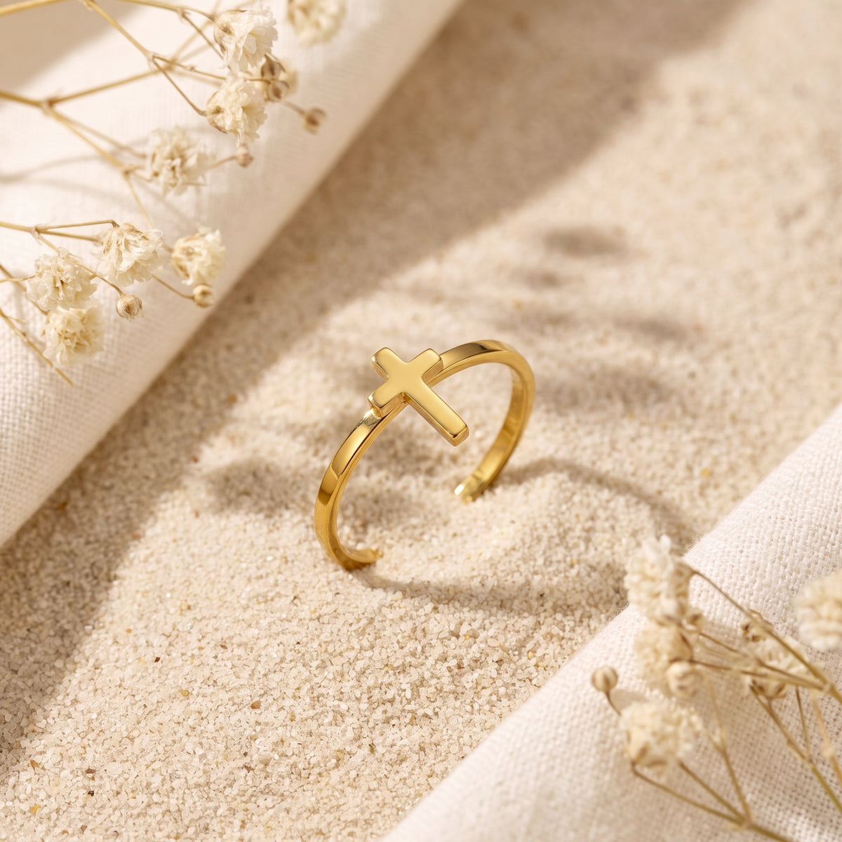 Gold-tone open cross ring standing on sand beside dried white flowers and neutral fabric.