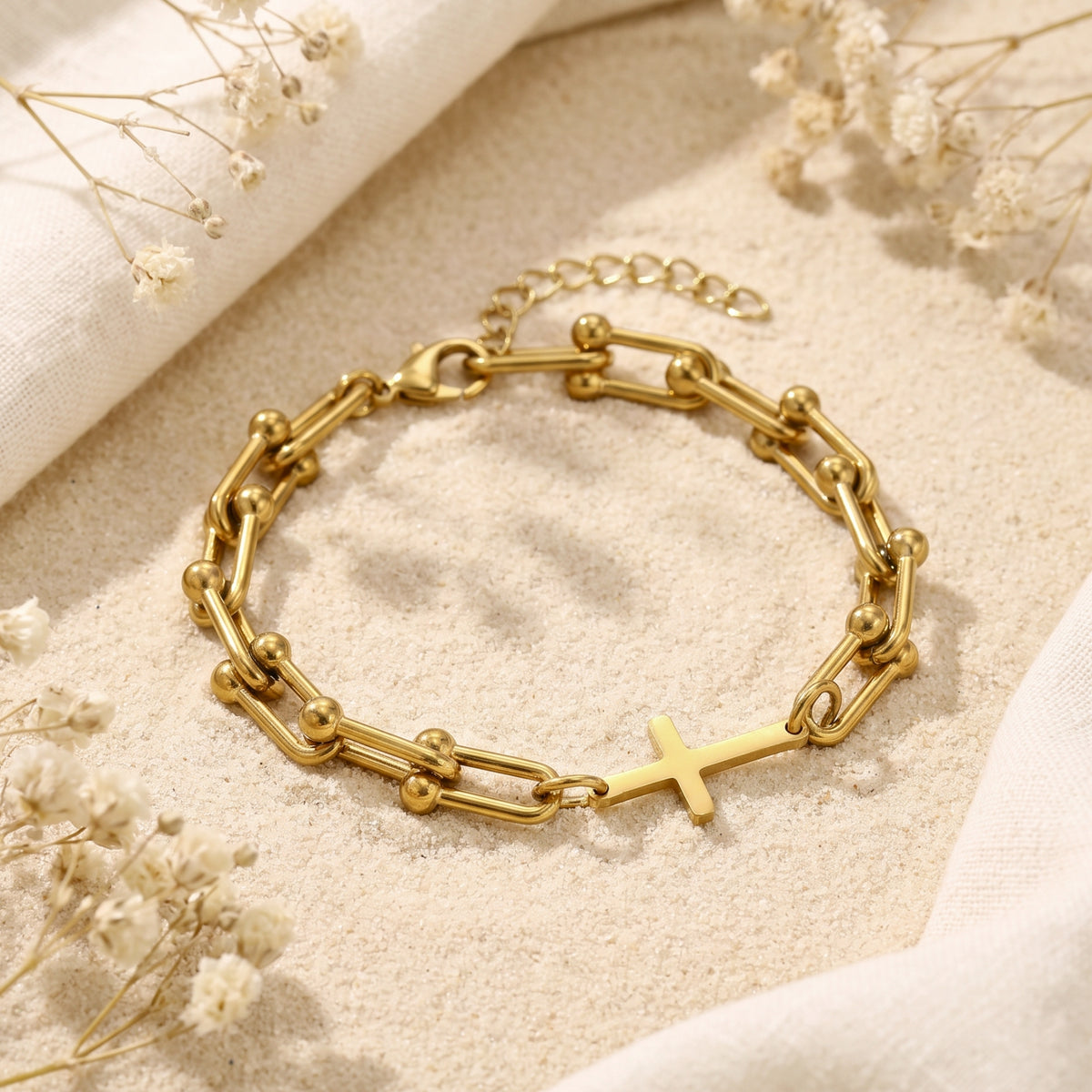 Gold-tone U-link bracelet with a horizontal cross centerpiece resting on sand among white dried flowers.
