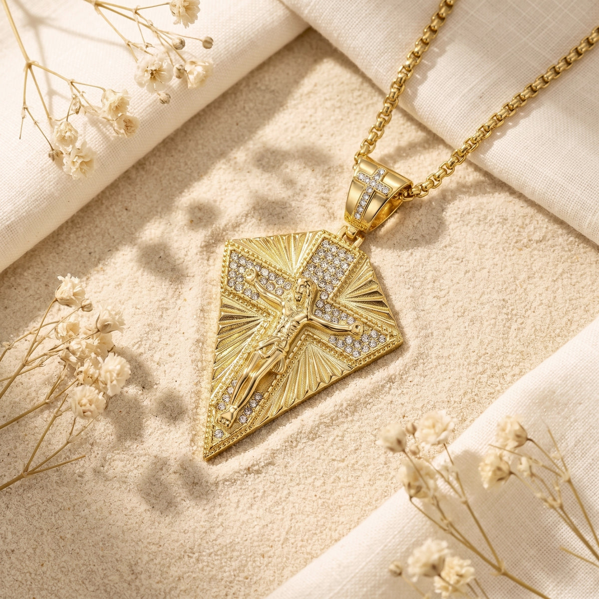 Gold-finished crucifix pendant necklace lying on sand among dried white flowers and folded fabric.