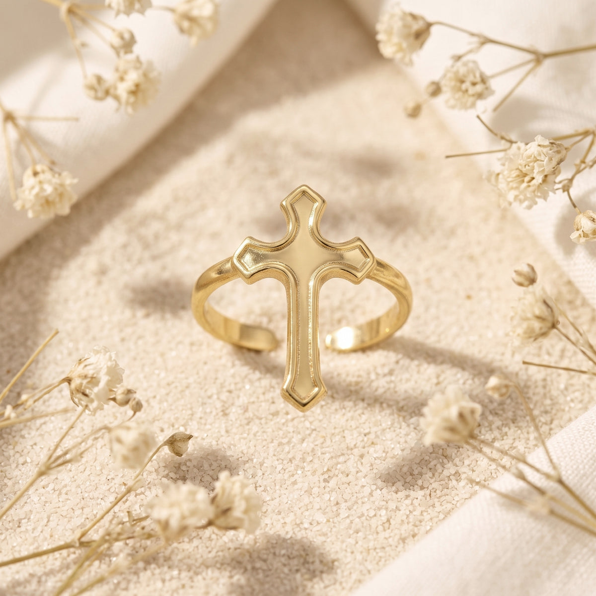 Gold-tone open ring with a contoured cross centerpiece displayed on fine sand with dried white flowers.