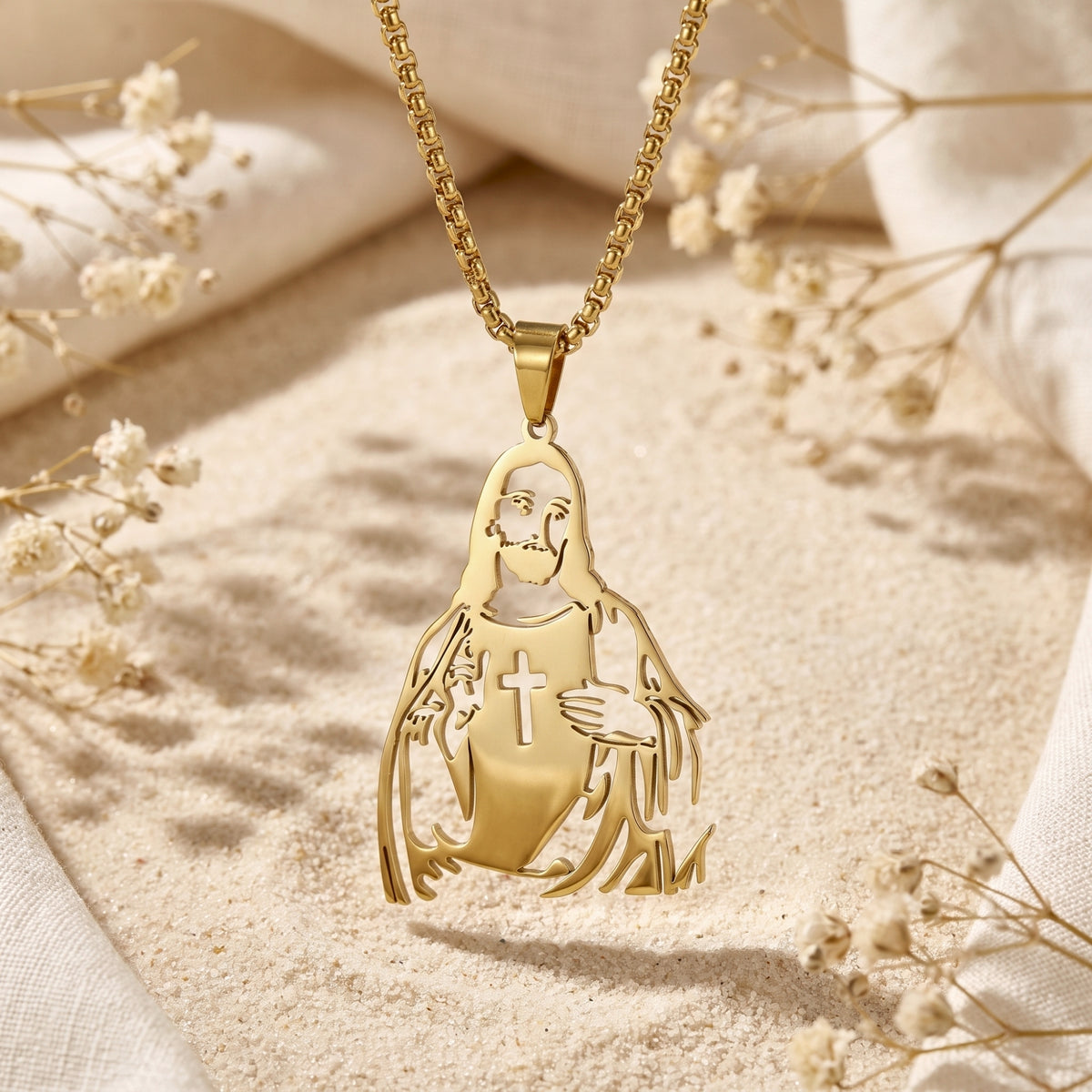 Gold-tone Christ silhouette pendant with a cross cutout hanging above sand, framed by dried white flowers.