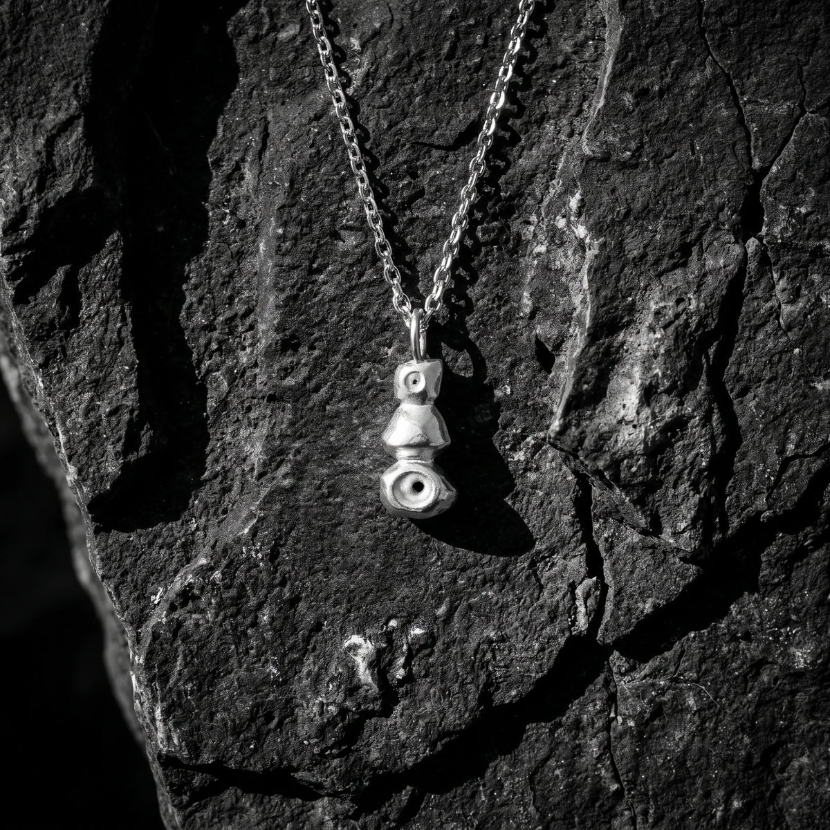 Silver cairn pendant with three stacked geometric forms hanging against dark textured rock.