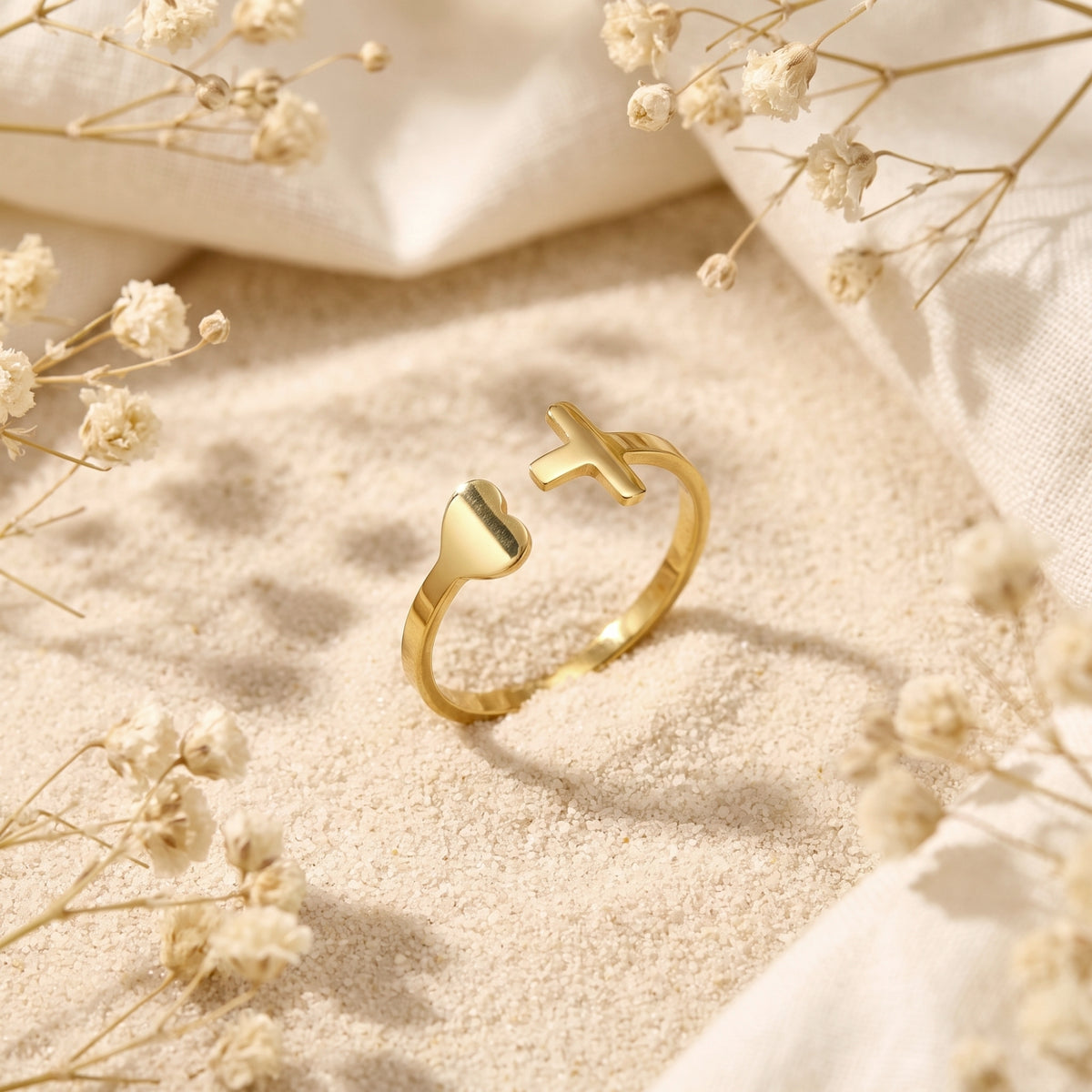 Gold-tone open ring with heart and cross motifs standing upright on white sand among dried white flowers.