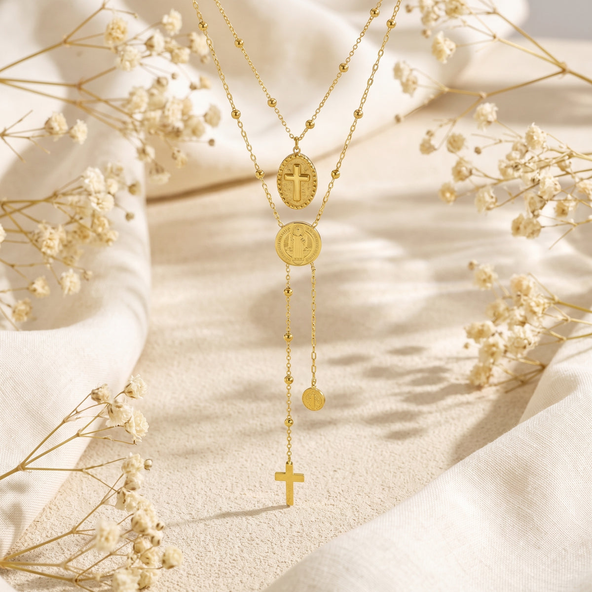 Gold-tone layered necklace with an oval cross medallion, round religious medal, and dangling cross on white fabric