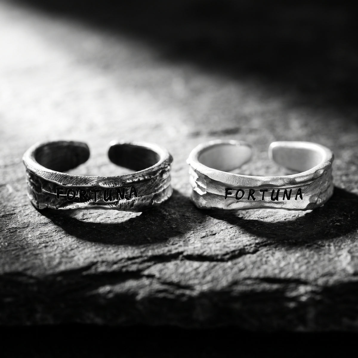 Two textured open silver rings stamped FORTUNA standing side by side on a dark textured surface.