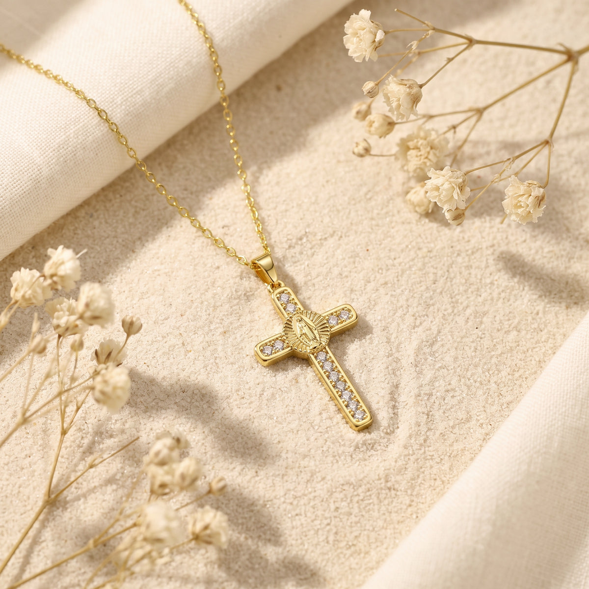 Gold-tone cross pendant necklace with clear stones resting on sand with dried white flowers and fabric.