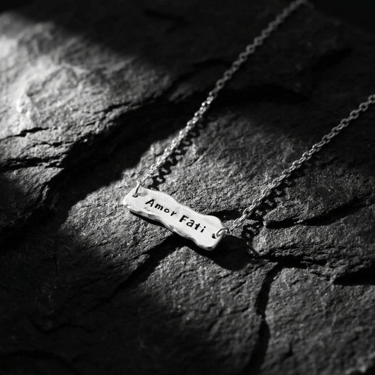 Amor Fati bar necklace arranged on dark textured slate, with the inscription visible on the pendant.