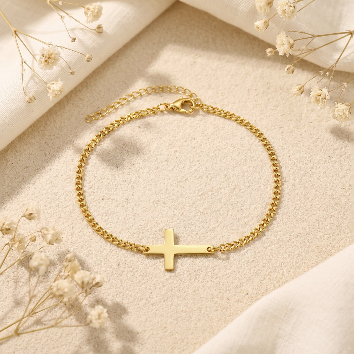 Gold-toned horizontal cross curb chain bracelet resting flat on sand, with white fabric and dried flowers around it.