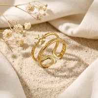 Gold-tone double-band cross ring with engraved script resting on sand among dried flowers and light fabric.