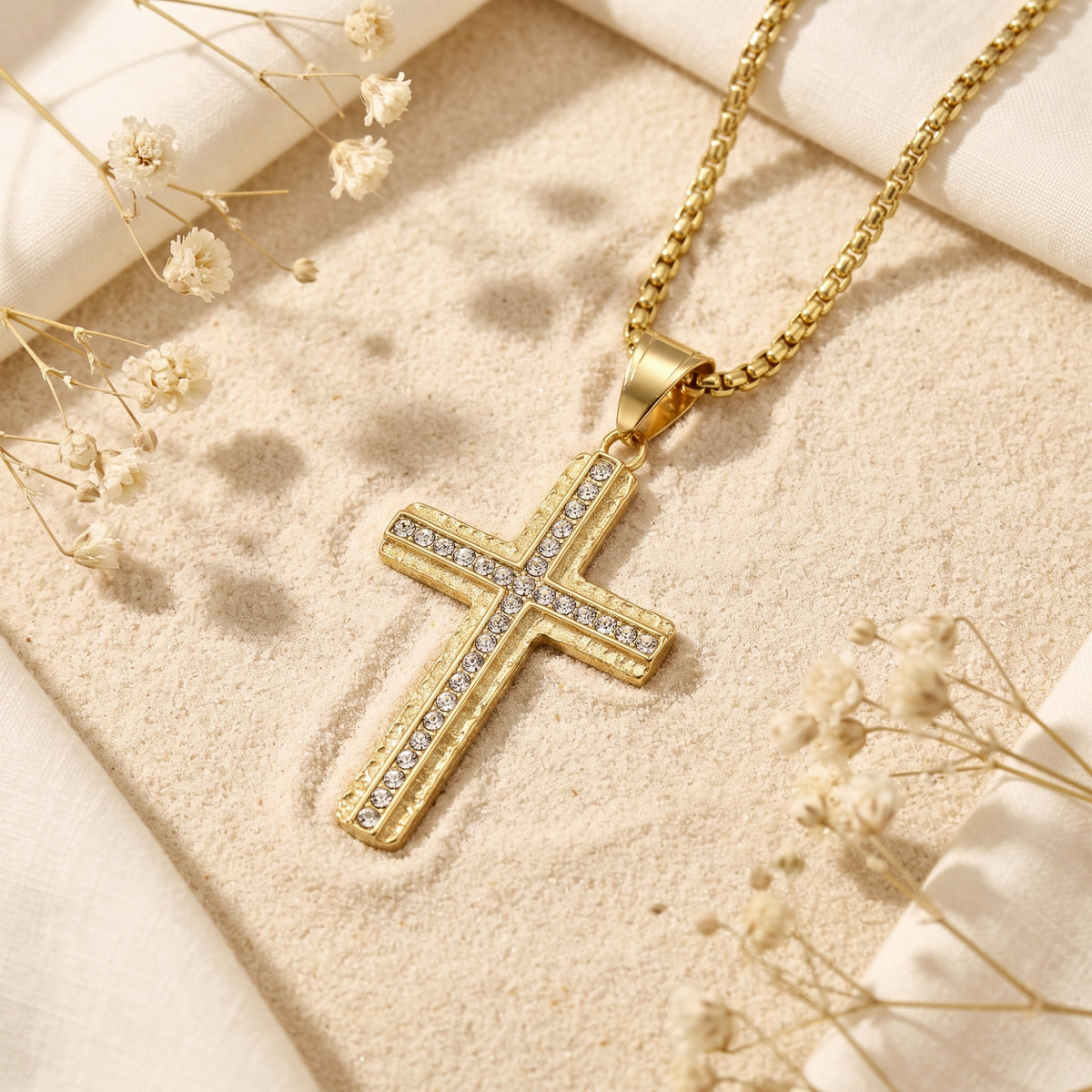 Gold-tone cross pendant with cubic zirconia inlay laid flat on sand beside dried white flowers.