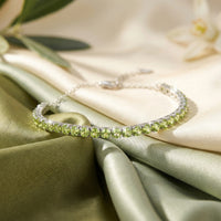 Round cut natural peridot tennis bracelet in sterling silver displayed on green and cream silk fabric.