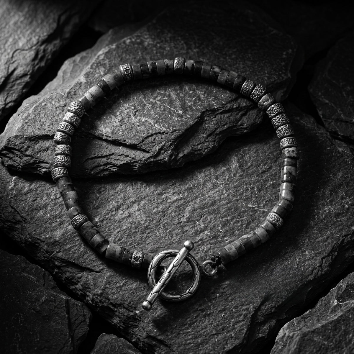 Overhead view of the silver toggle bracelet in a circular loop on dark slate, showing etched cylinder beads and clasp.