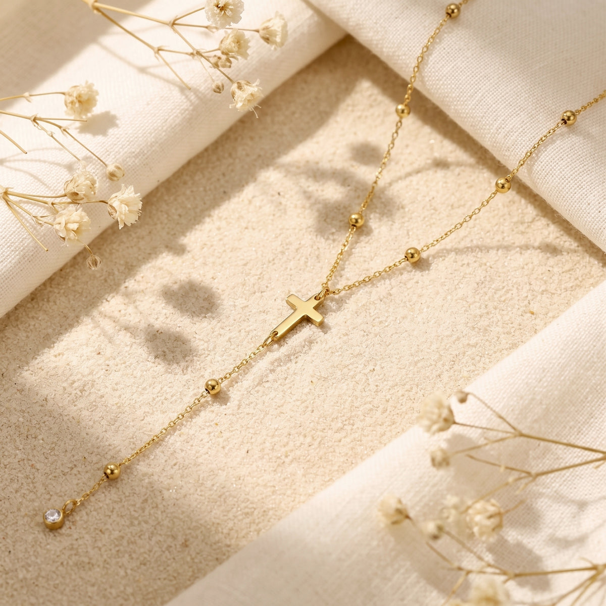Gold-tone cross lariat necklace lying diagonally on sand, with bead stations and a cubic zirconia drop