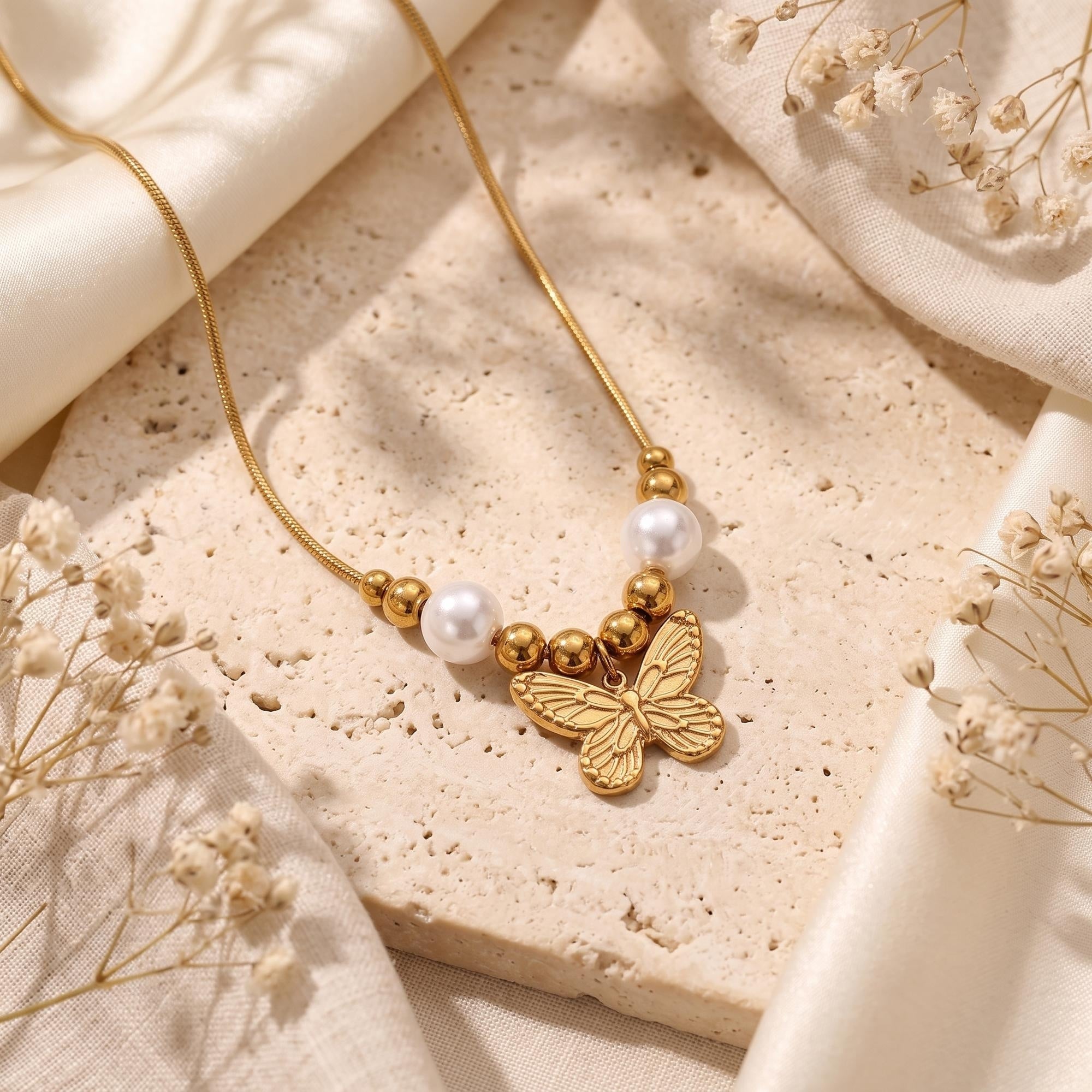 Gold-tone butterfly pendant necklace with two white pearls and metal beads on a textured stone tile.