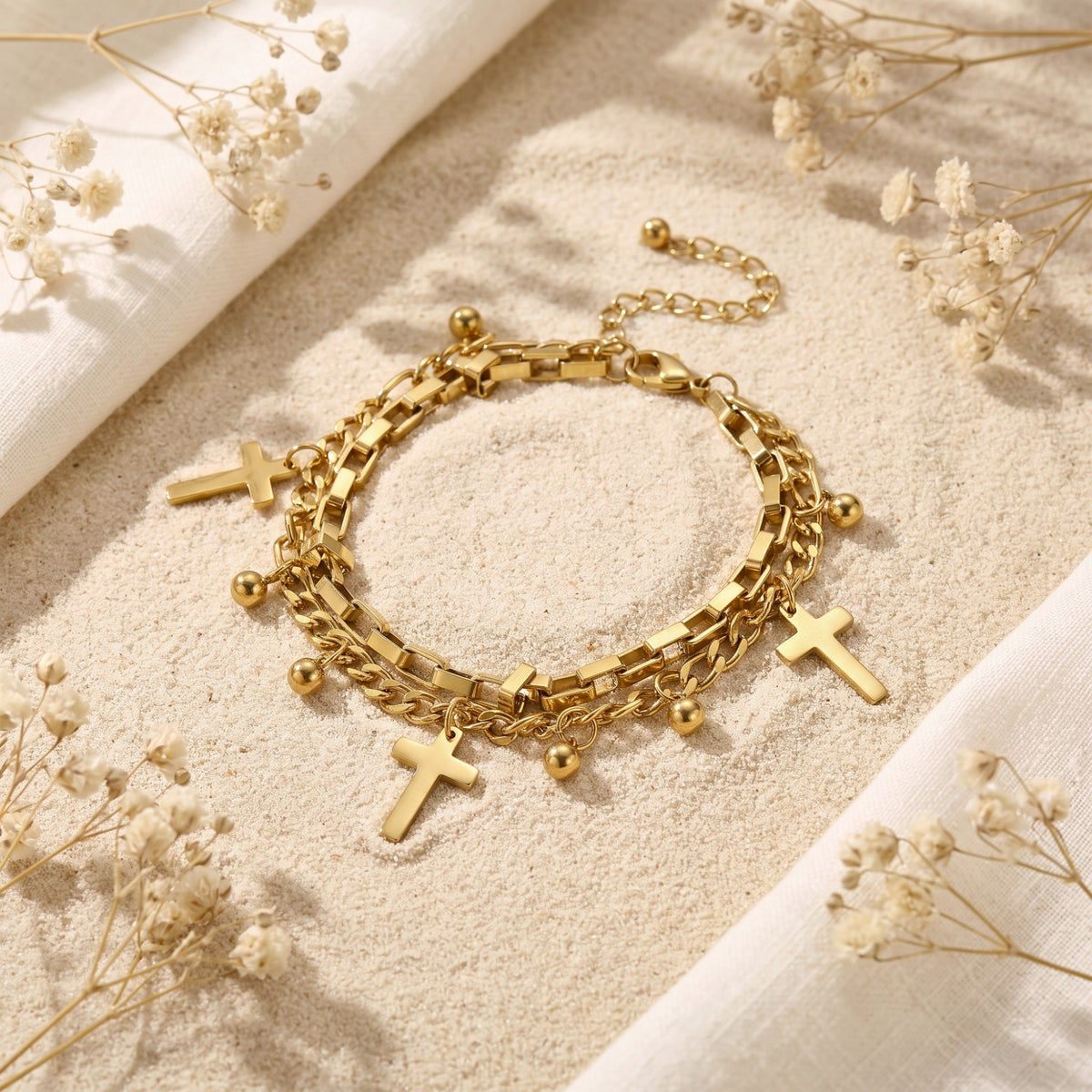 Gold-tone double-layer bracelet with cross and bead charms laid flat on sand among white dried flowers.