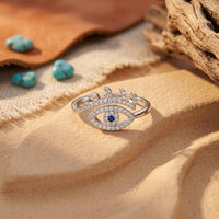 Sterling silver eye-shaped ring with a blue center stone on sand with turquoise stones in the background.