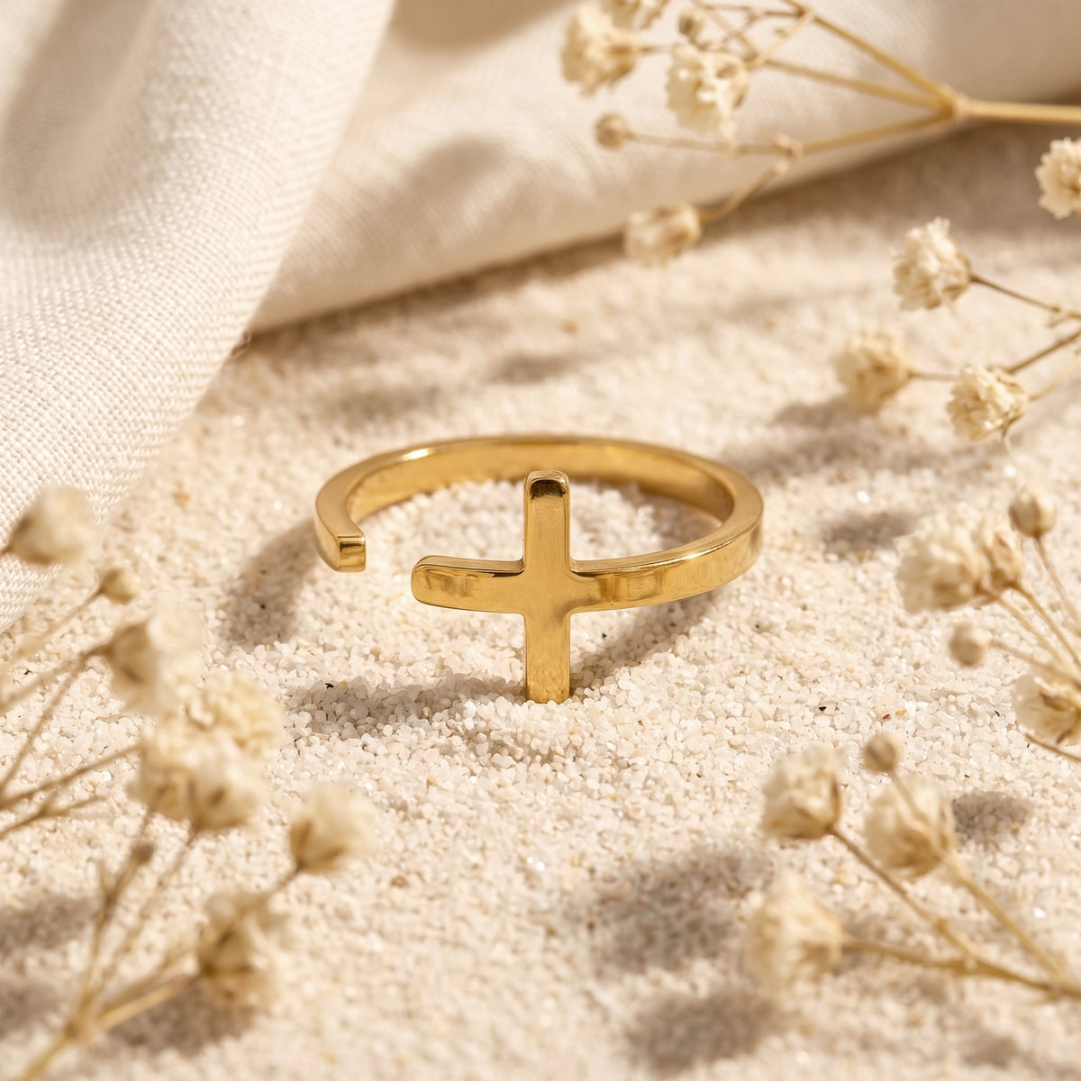 Gold-tone cross open ring resting upright in fine white sand with dried white flowers.