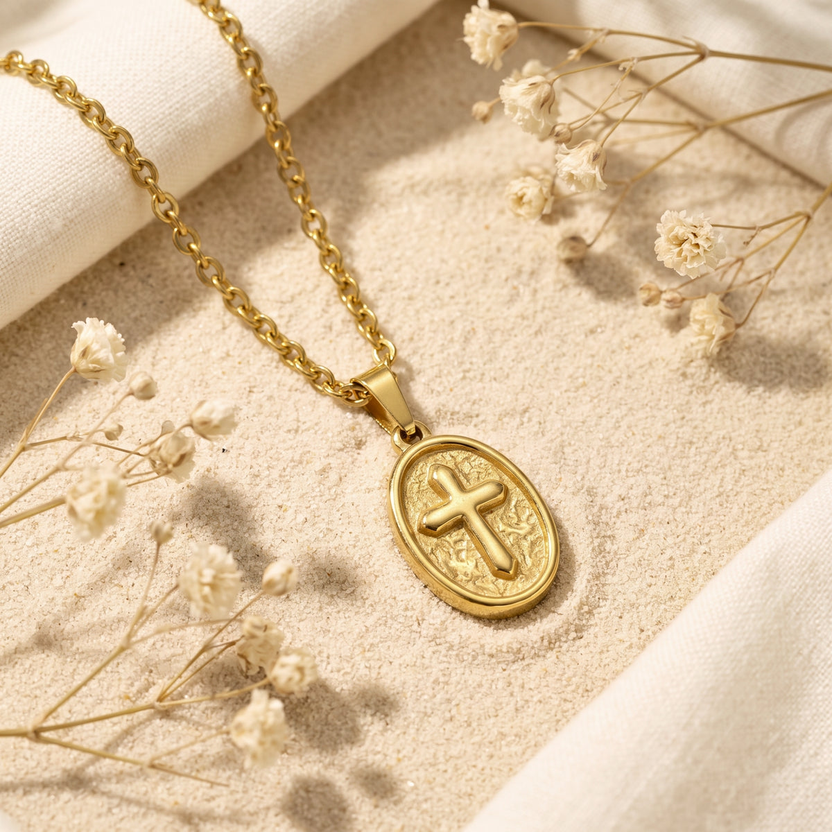 Gold-tone oval cross medallion necklace lying diagonally on sand with dried white flowers and linen.