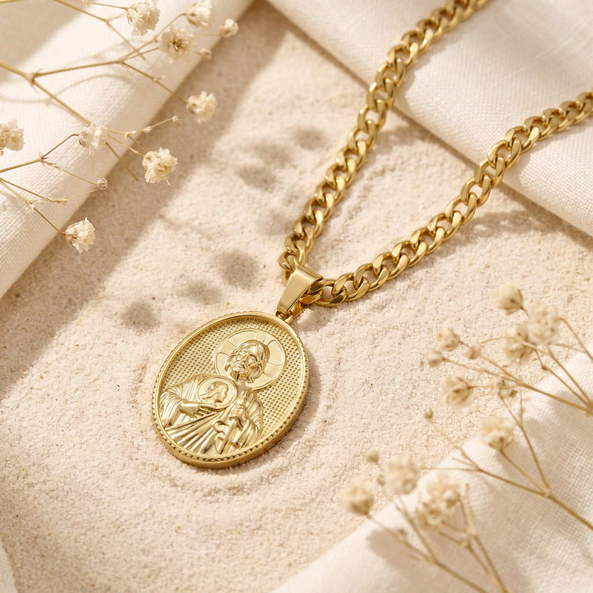 Gold-tone oval Saint Jude pendant necklace lying on sand with white fabric and dried flowers.