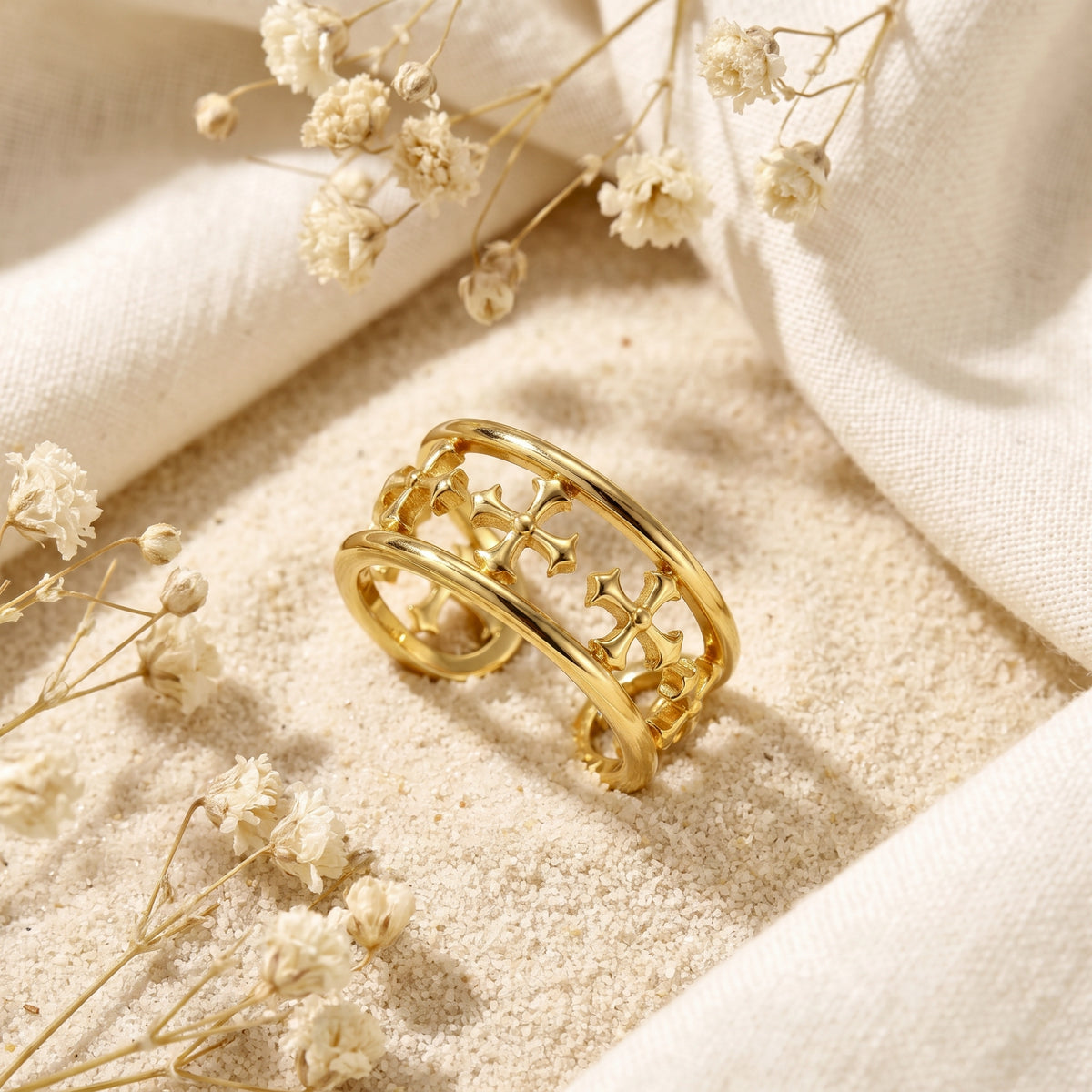 Gold-tone double-rail cross cuff ring resting on fine sand beside white dried flowers.