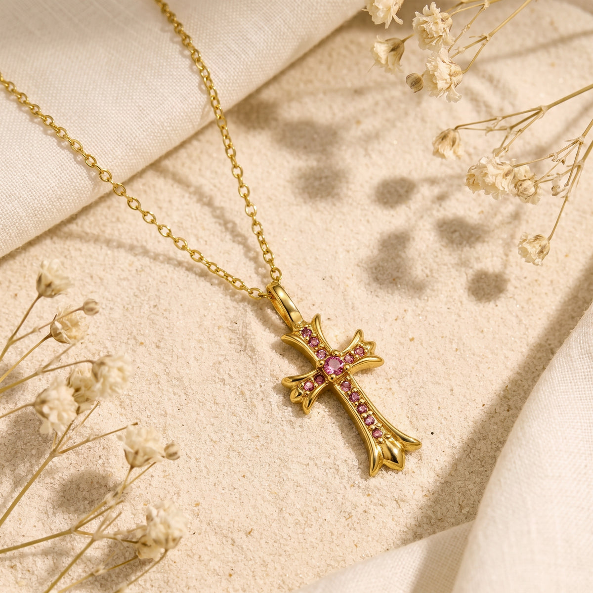 Gold-tone cross necklace with pink accents laid diagonally on sand beside white dried flowers.