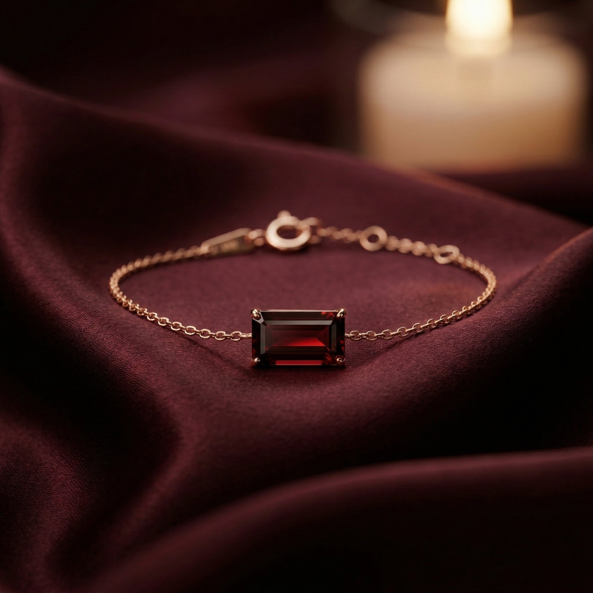 Emerald-cut natural garnet bracelet in 18K rose gold displayed on burgundy silk with a soft candlelit background.