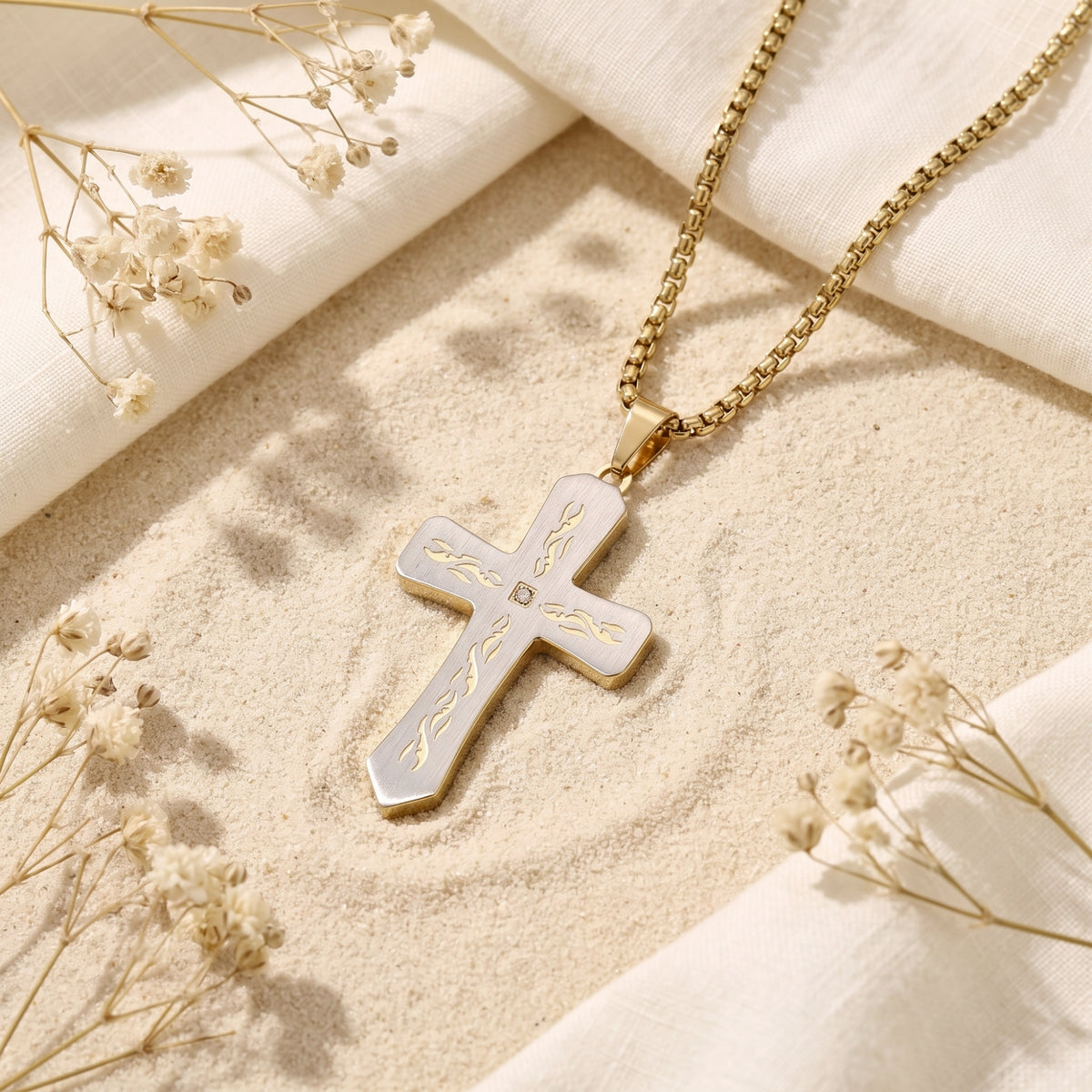 Two-tone cross pendant and gold-tone box chain lying diagonally on sand beside dried flowers and folded white fabric.