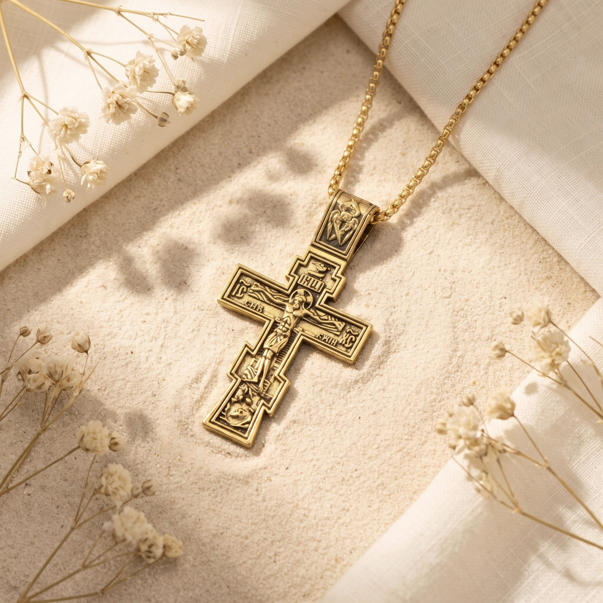 Gold-tone Eastern cross pendant and chain lying on pale sand among white fabric and dried flowers.