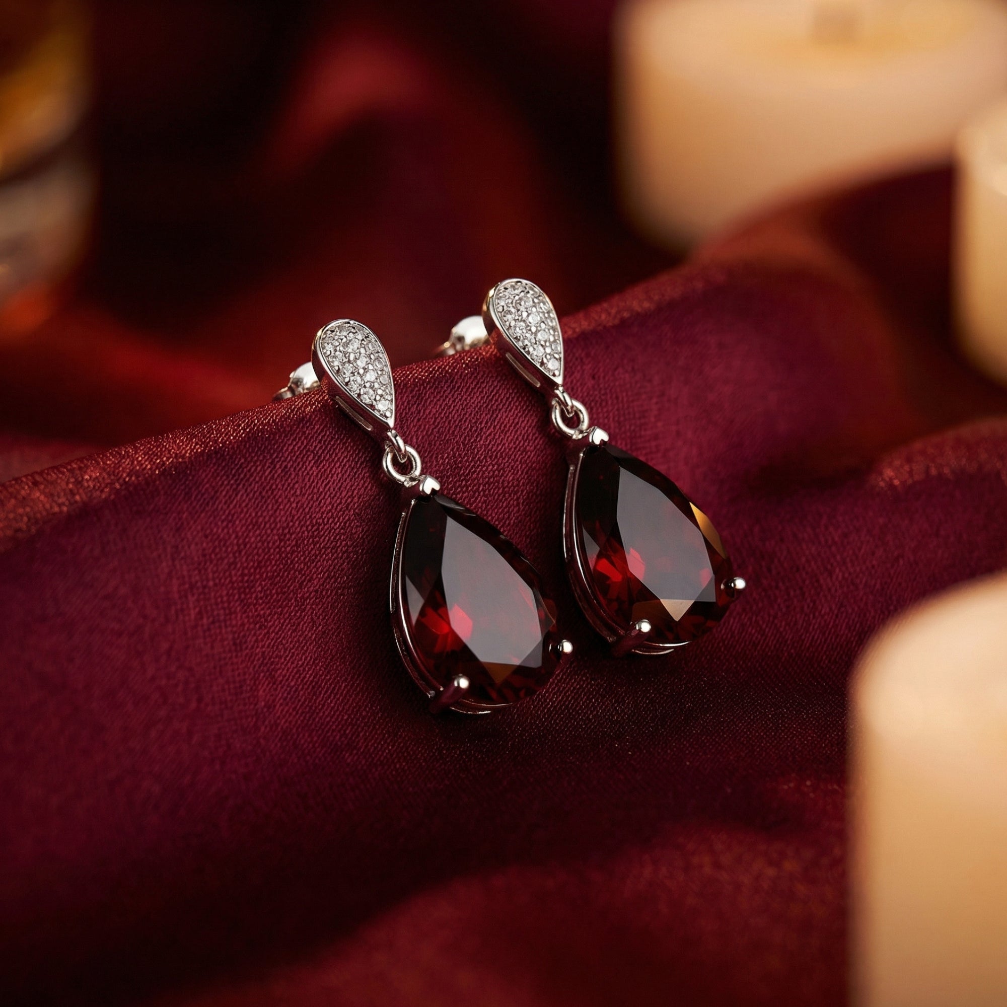Pear cut garnet drop earrings in sterling silver resting on elegant burgundy silk fabric with soft candle lighting.